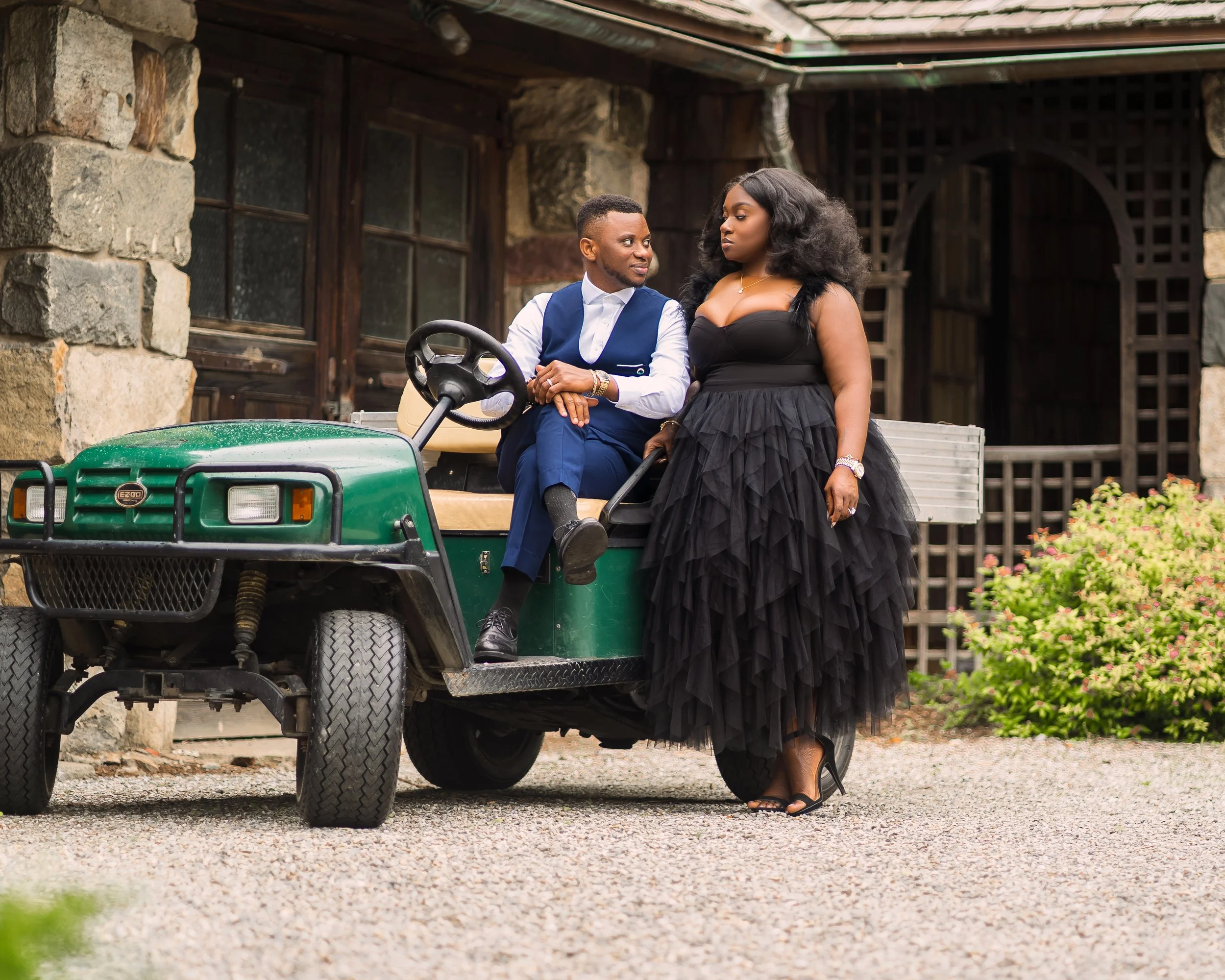A man and woman sitting on the back of a green golf cart parked outside a rustic building with stone walls and wooden windows. The woman in a black dress and high heels is standing next to the man, who is wearing a suit vest and white shirt.