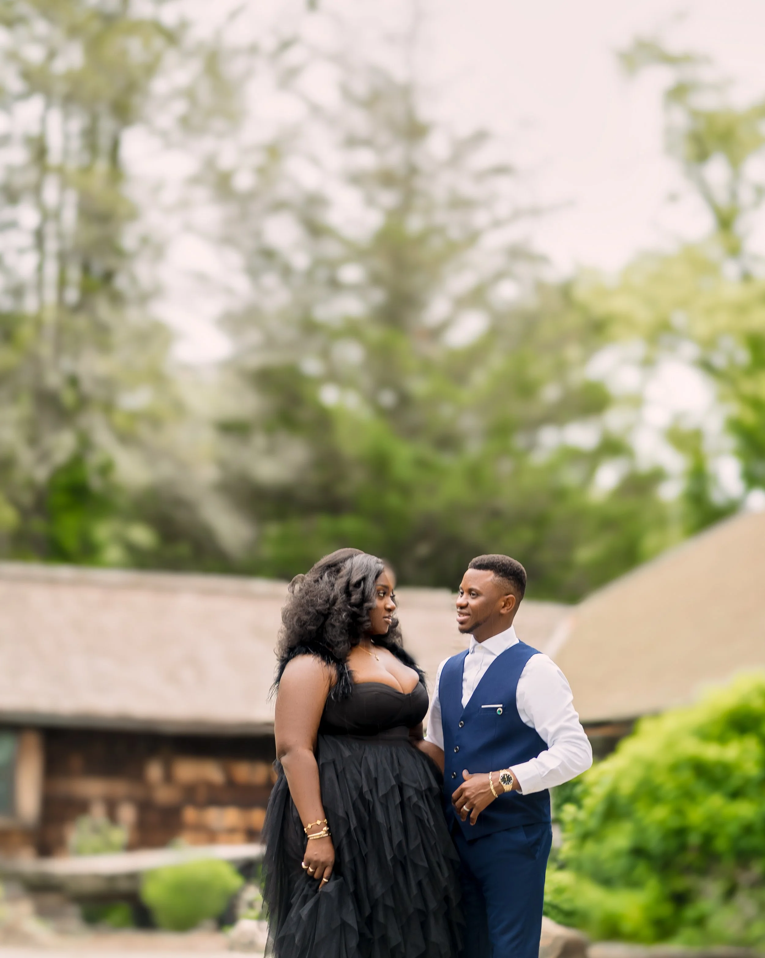 A couple standing close together outdoors; the woman is wearing a strapless black dress with a tulle skirt and gold jewelry, and the man is wearing a blue suit vest over a white shirt with a gold watch; they are in a green park with trees and a woode