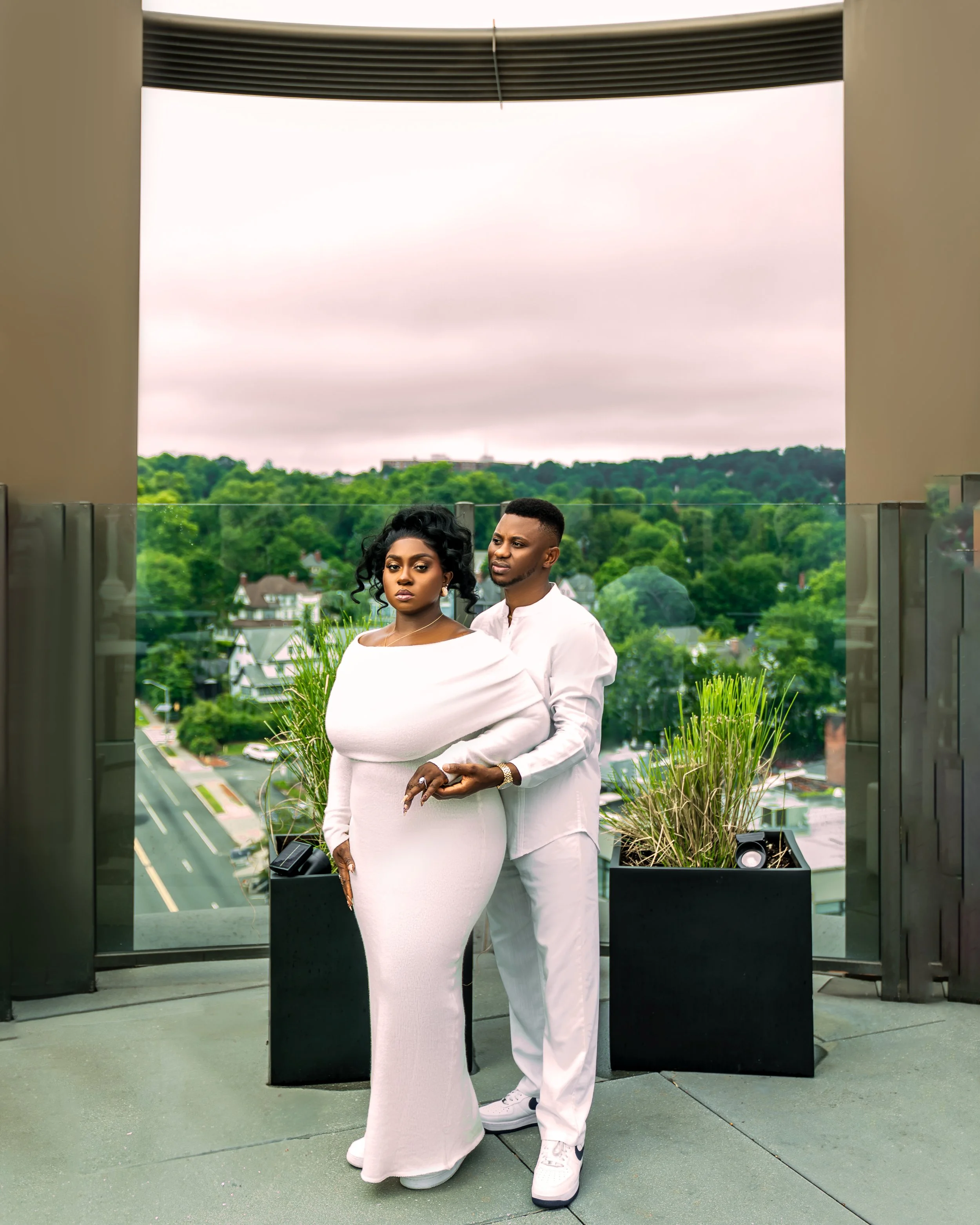 A couple dressed in white standing outdoors on a rooftop or balcony with a scenic view of green trees and houses in the background, under a cloudy sky.