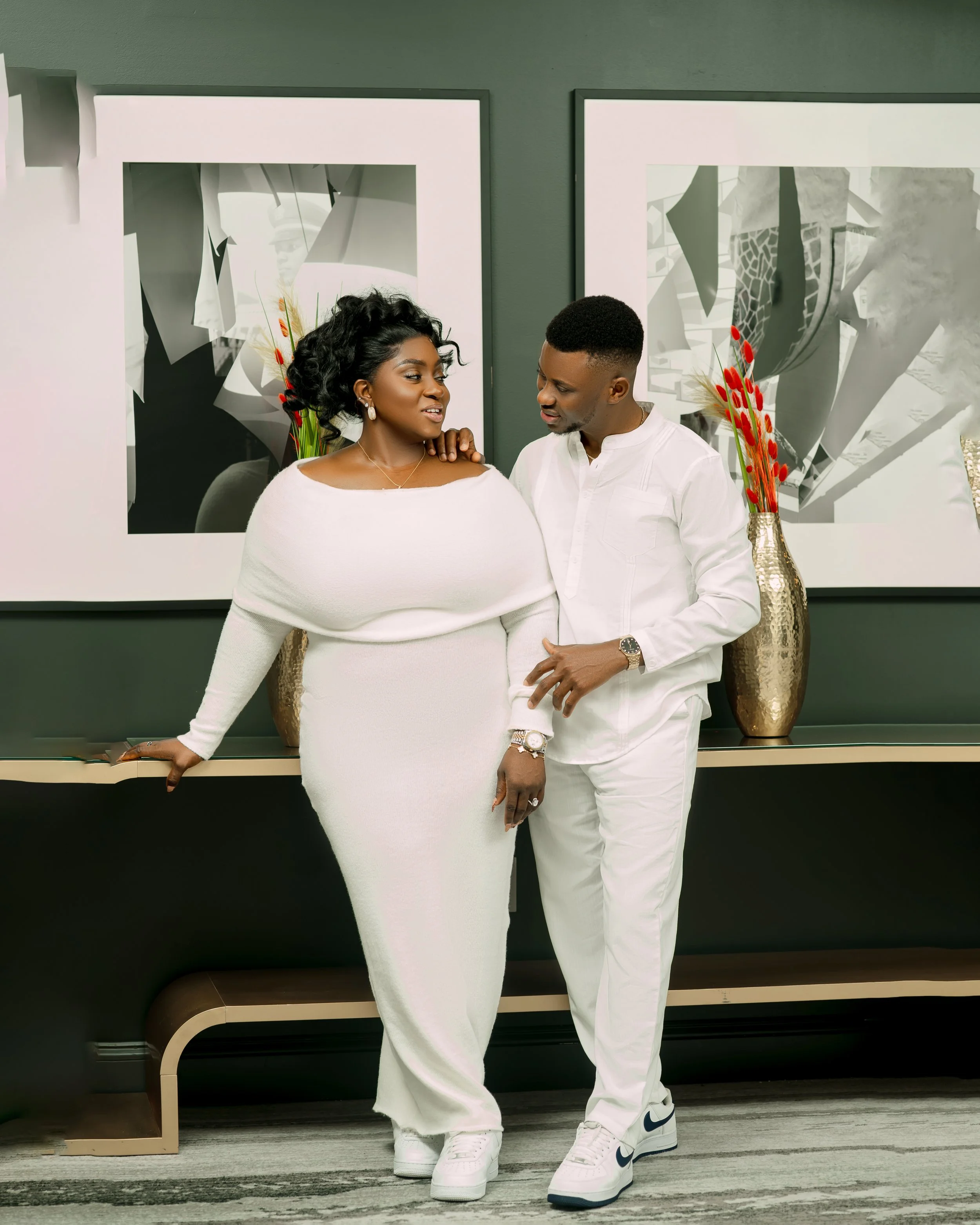 A black couple dressed in white stands close together in front of a modern grey and white wall with twin mirrors and floral decor. The woman is wearing a long white dress with off-the-shoulder neckline, pearl earrings, and a gold necklace, while the 