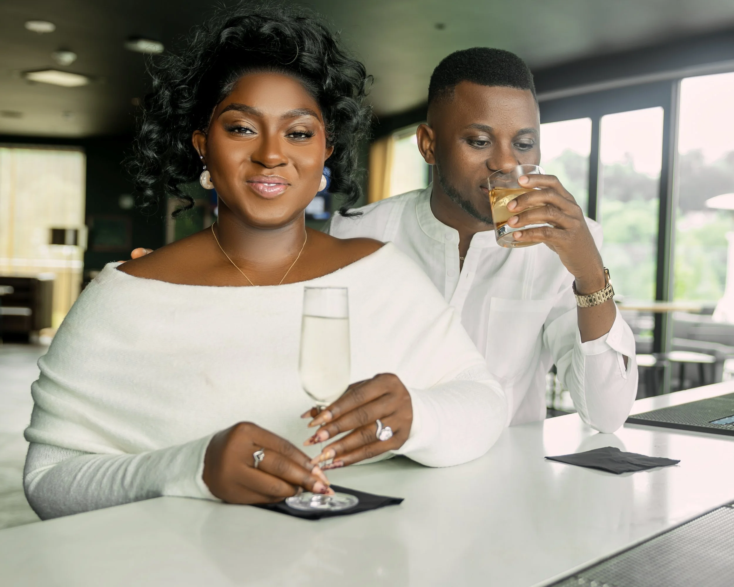 A woman with black, curly hair and a man with a short, flat-top haircut sitting at a table. The woman is smiling gently while the man is drinking a glass of what appears to be white wine or champagne. The setting is modern with large windows in the b