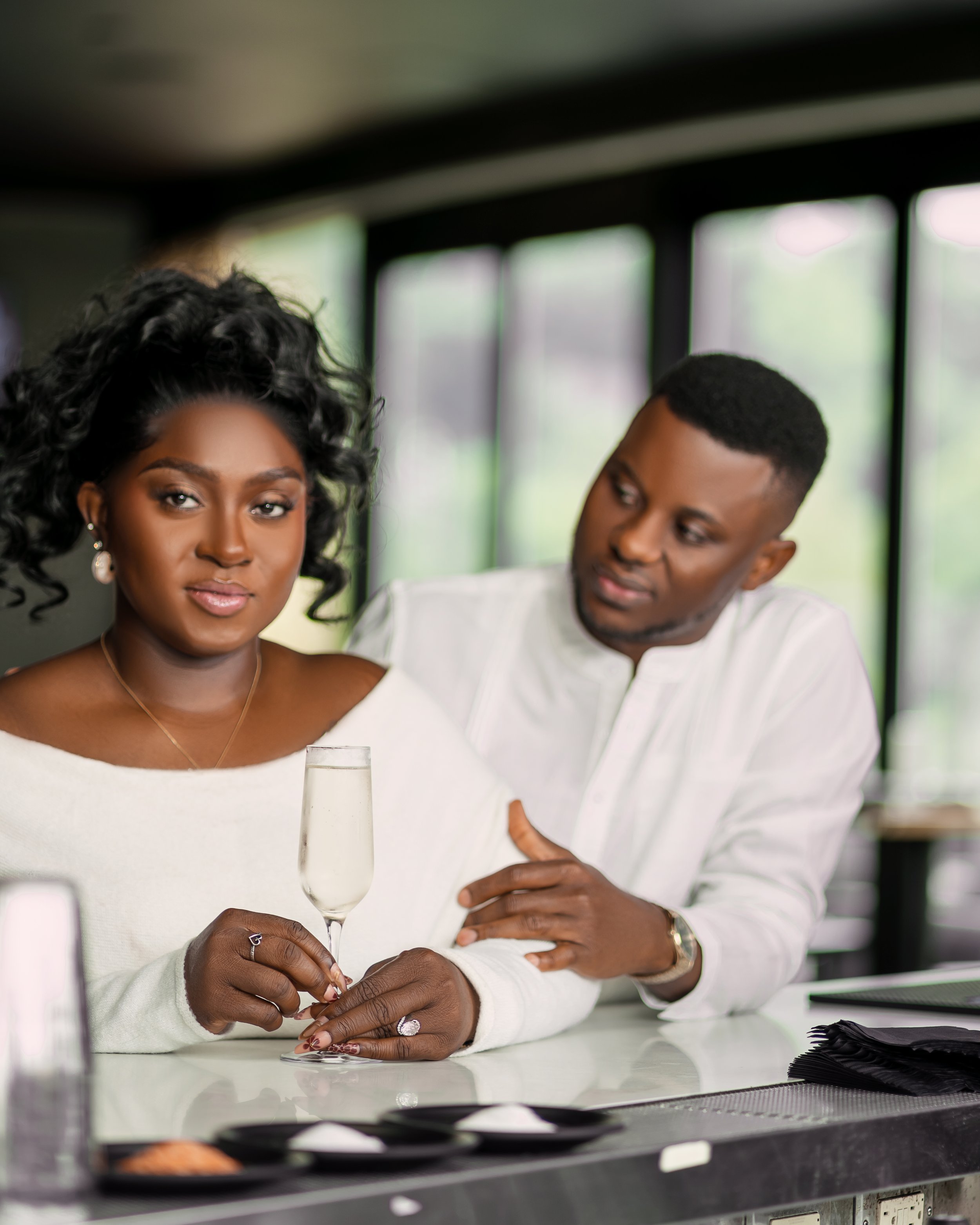 A woman is sitting at a counter with a man beside her, filling out a form or writing on a paper, holding a glass of sparkling wine or champagne, and they are in front of a window with blurred outdoor scenery.