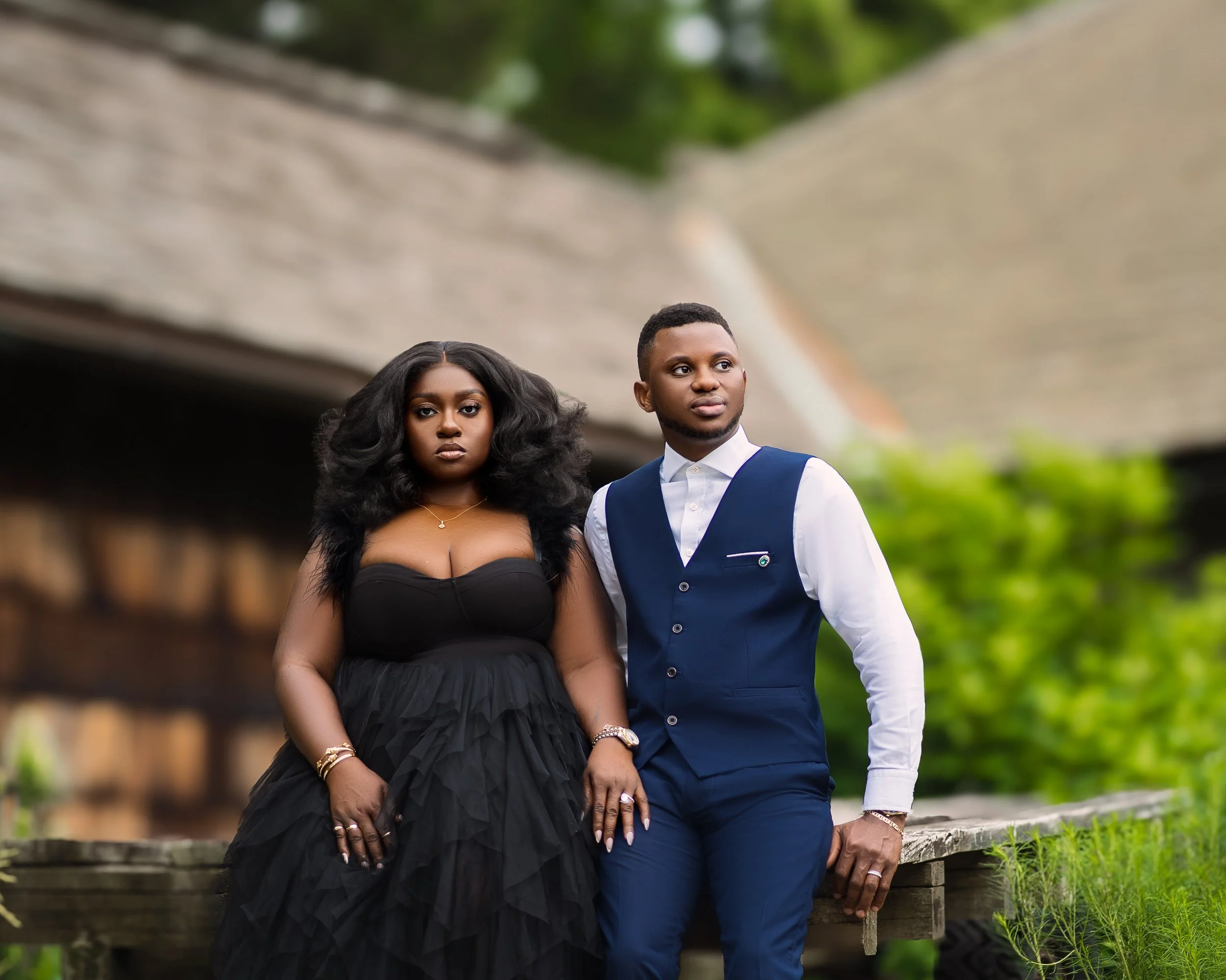 A man and a woman dressed formally standing outdoors near a wooden bridge with greenery in the background.