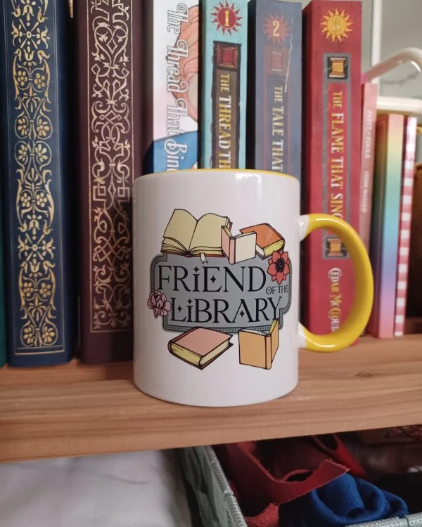 A photo of a ceramic mug sitting on a bookshelf. The mug is white with a yellow handle, the design on it the words "Friend of the Library" surrounded by flowers and books.