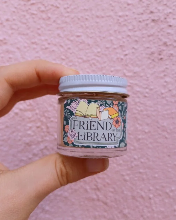  A photo of a white hand holding a miniature glass jar candle in front of a pink wall. The candle is labeled "Friend of the Library." 