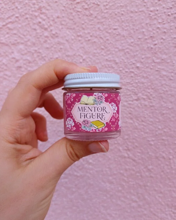  A photo of a white hand holding a miniature glass jar candle in front of a pink wall. The candle is labeled "Mentor Figure." 