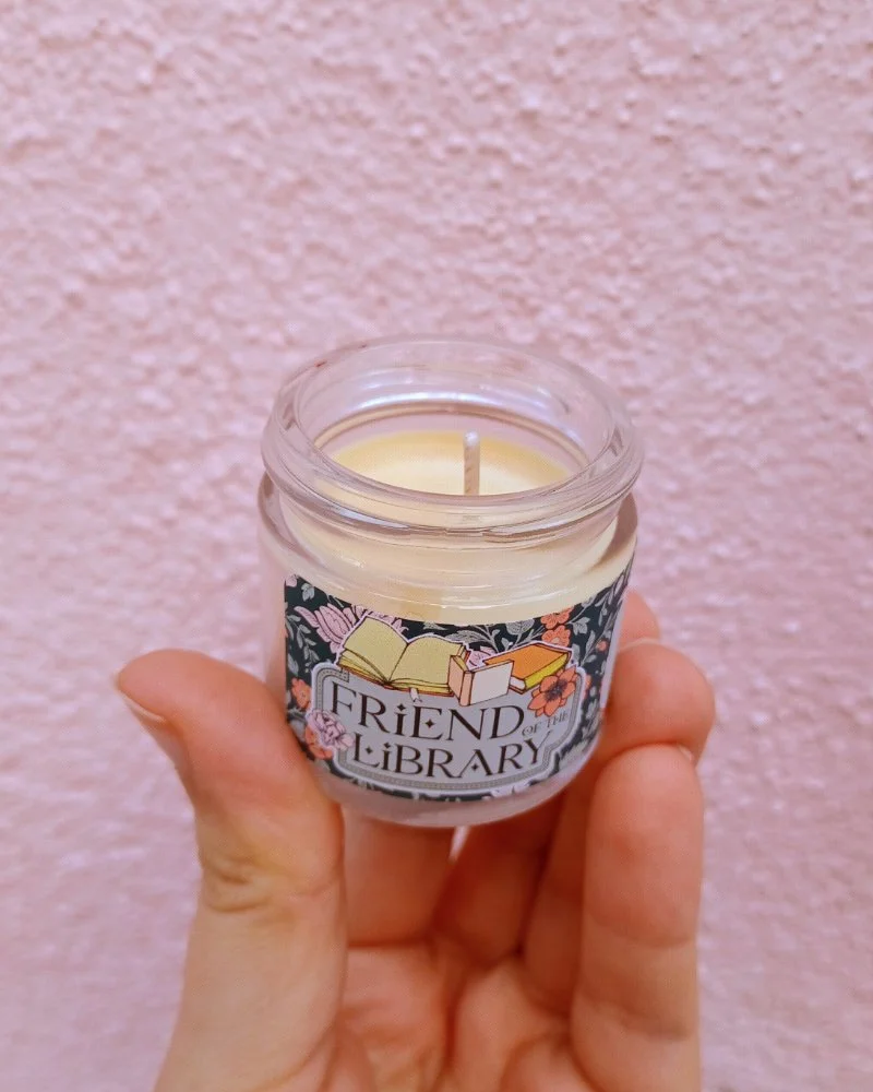  A photo of a white hand holding a miniature glass jar candle in front of a pink wall. The candle is tilted to show the wax inside. 