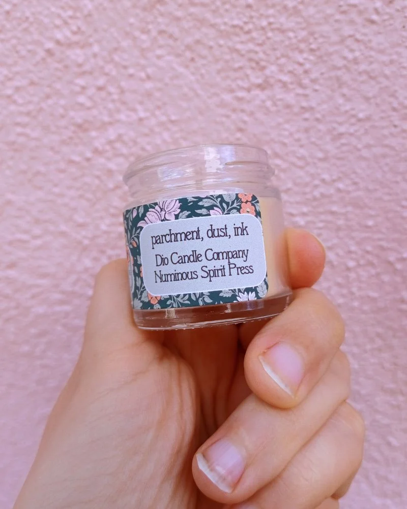  A photo of a white hand holding a miniature glass jar candle in front of a pink wall. The candle scent is labeled “parchment, dust, ink.” 