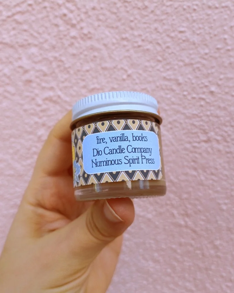  A photo of a white hand holding a miniature glass jar candle in front of a pink wall. The candle scent is labeled “fire, vanilla, books” 