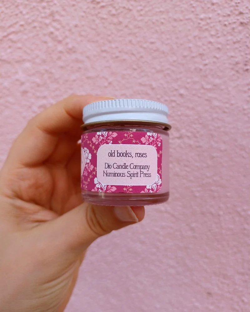  A photo of a white hand holding a miniature glass jar candle in front of a pink wall. The candle scent is labeled “old books and rose” 