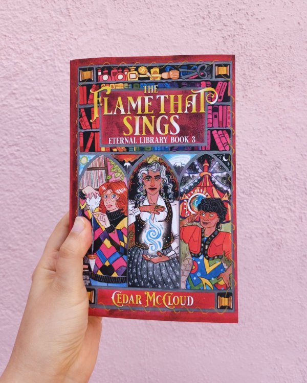 A photo of a white hand holding a paperback book in front of a pink wall. The book is THE FLAME THAT SINGS by Cedar McCloud.