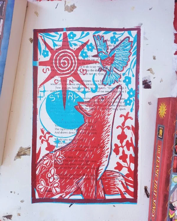 A photo of a handmade linocut print surrounded by books. The print features a red fox and bluebird with flowers on top of a book page pasted to handmade recycled paper with flower petals in it