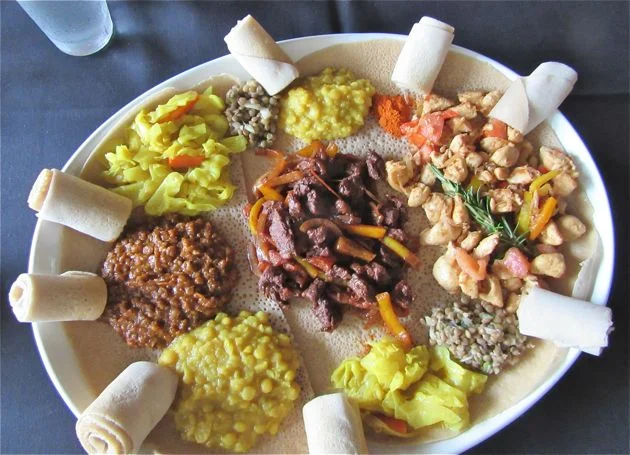 A feast of meat and vegetable tibs and wots served atop injera bread with rolls of injera for grabbing morsels of food.