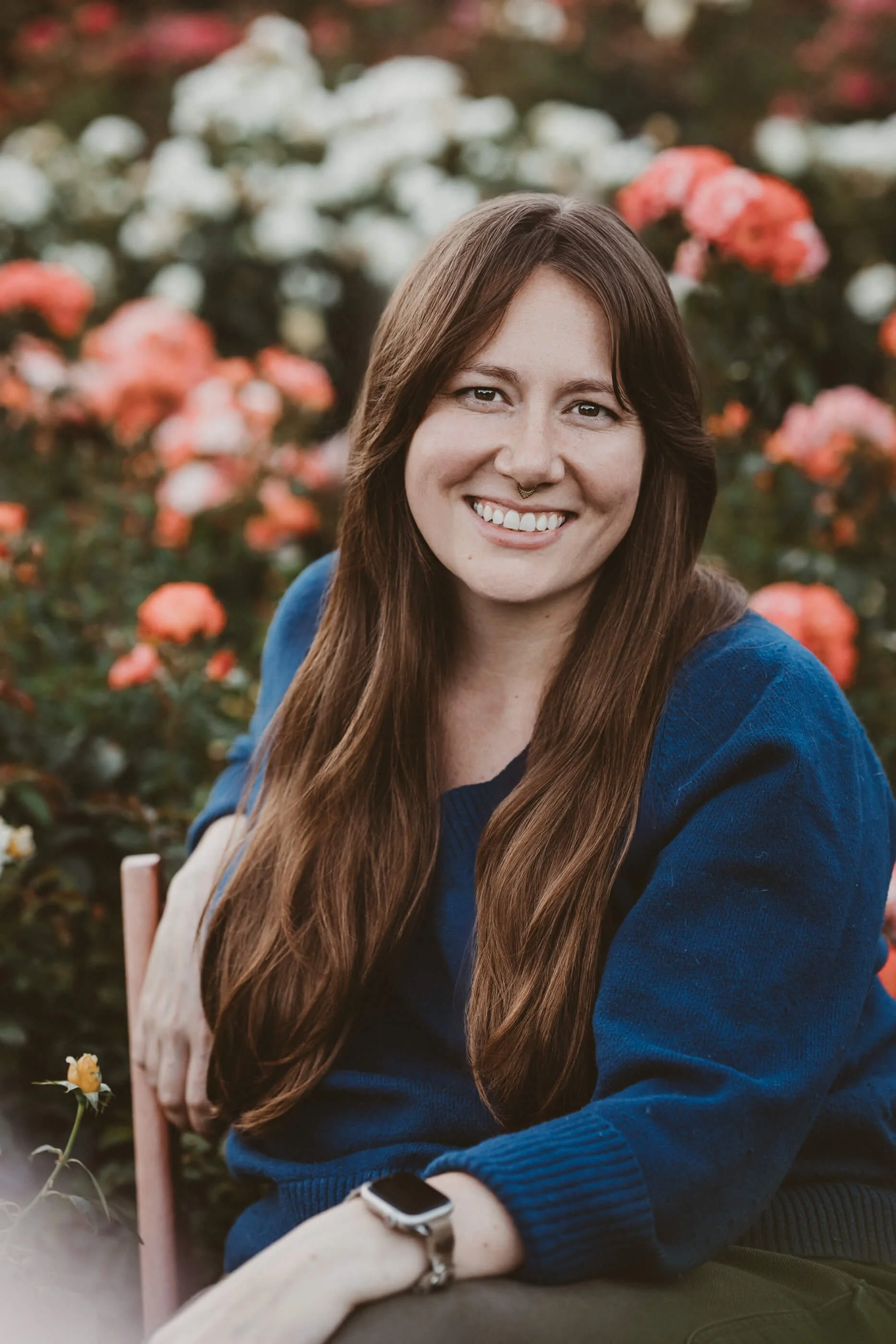 Portland doula Elle sits and smiles warmly at the camera with blooming roses behind her.