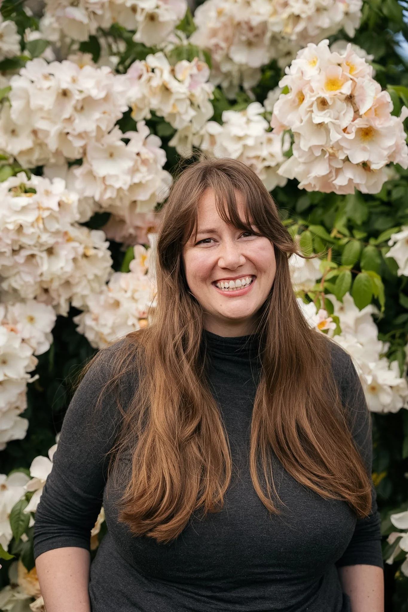 Birth Doula Portland Elle Dieal stands smiling warmly in front of a bloom of light pink and white flowers.
