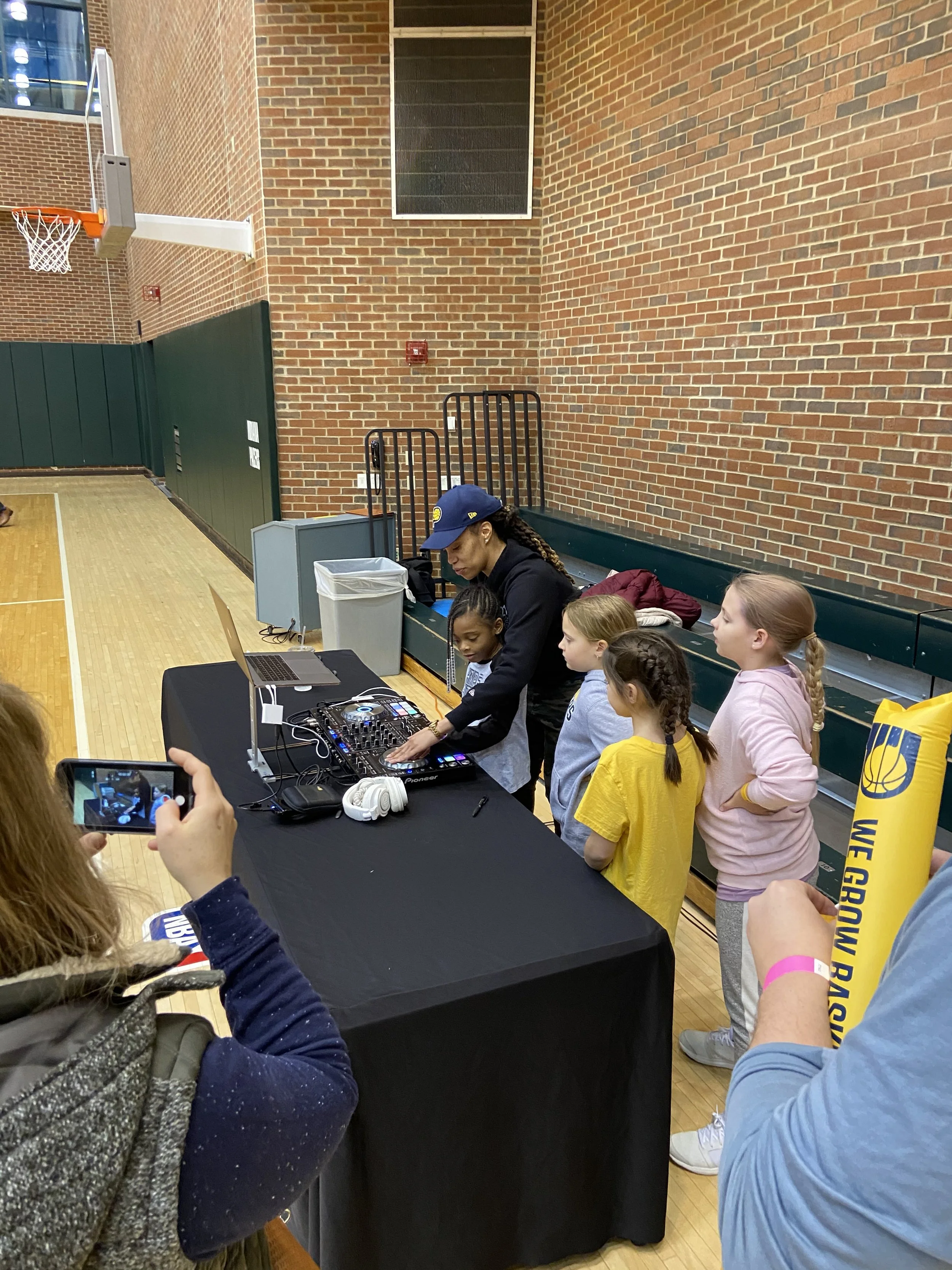 A woman DJ appears to be performing at a school gymnasium, surrounded by children who are watching her and taking pictures. The DJ is wearing a blue cap and black clothing, with a sound mixer and laptop in front of her. The gym has a brick wall, a basketball hoop, and some gym equipment.