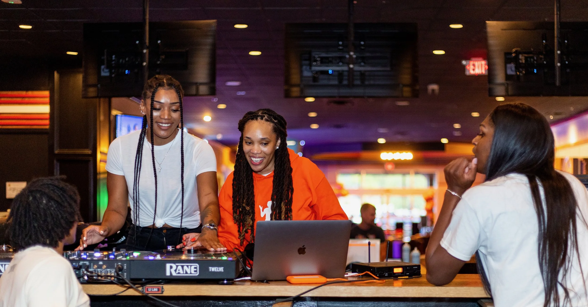 Three women and one man are gathered around a DJ controller and laptop, smiling and engaging in conversation in a dimly lit indoor space, possibly a bar or club.