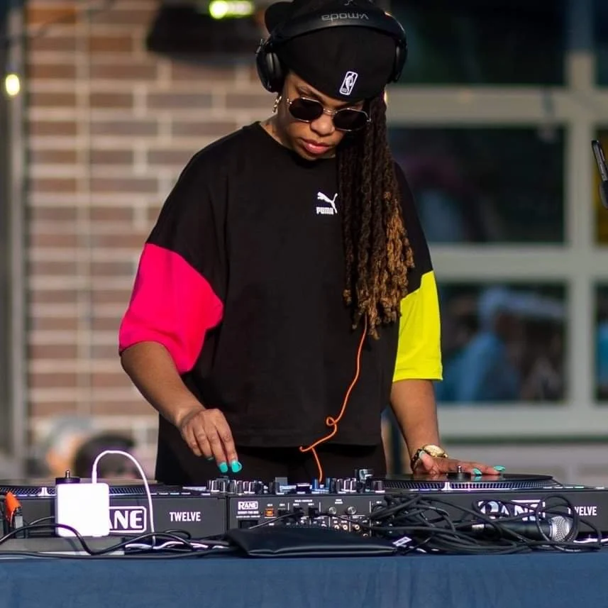 A woman DJ mixing music outdoors, wearing sunglasses, a black cap, a black and colorful Puma shirt, and headphones.