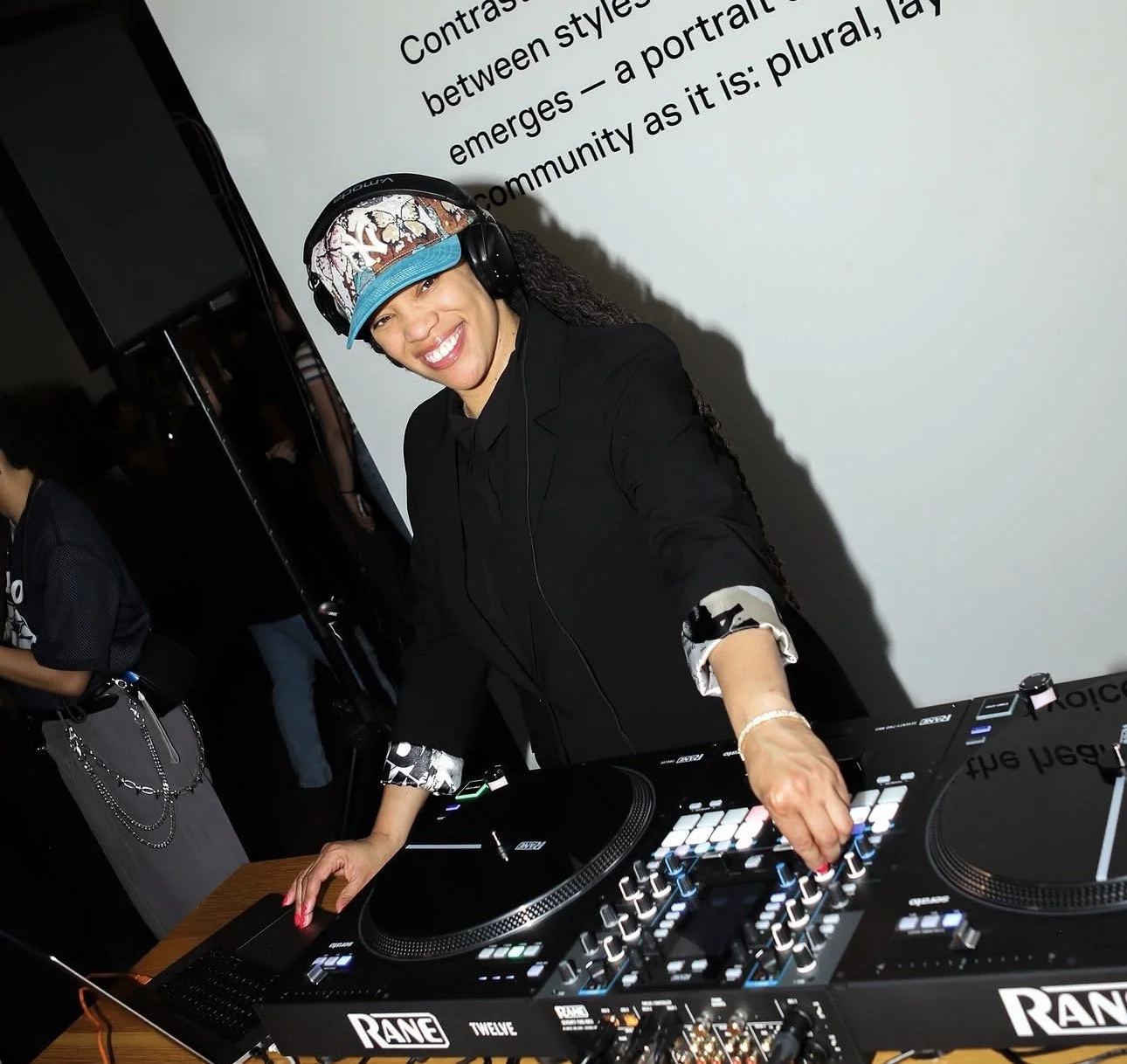 A woman in a black suit and hat with a colorful band, wearing headphones, smiling while DJing at a mixing console. She is in a dimly lit room with a white wall and partial text reading "Contr... between styles" in the background.