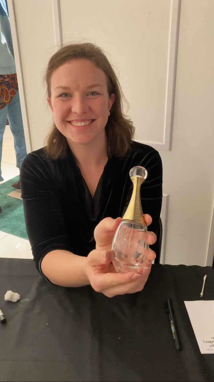 A smiling woman with shoulder-length brown hair holding a clear glass perfume bottle with a gold-colored cap shaped like a cone, in an indoor setting.
