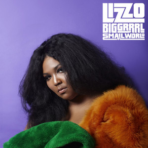A woman with long, voluminous black curly hair against a purple background, wearing a fur coat with green and orange sections, looking at the camera.