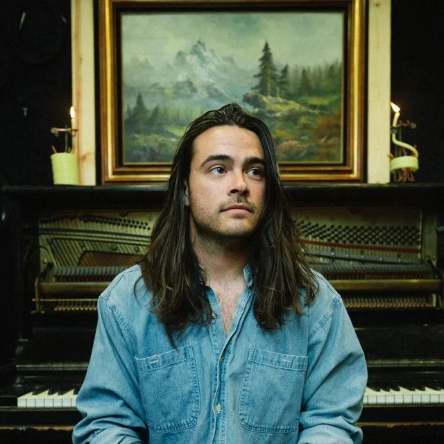 A man with long dark hair and a denim shirt sitting in front of a piano with a landscape painting behind him.
