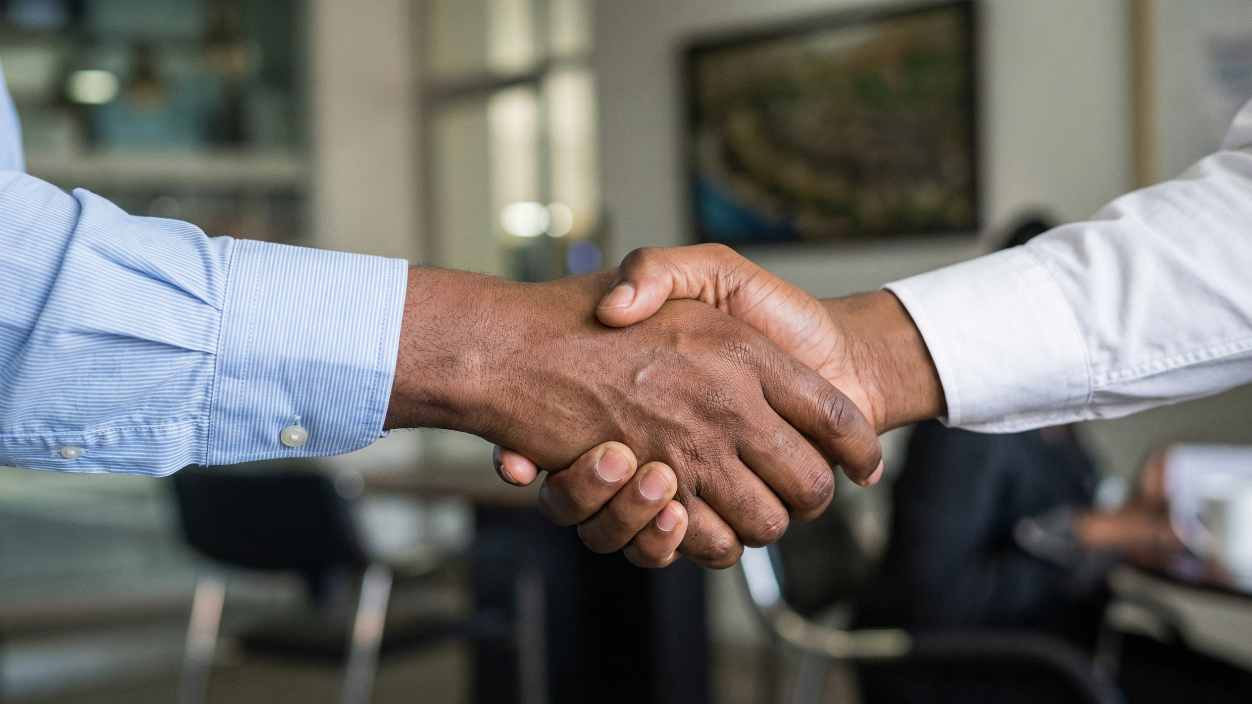  A close-up, medium shot of a handshake between two individuals with dark skin tones. One person wears a long-sleeved, light blue striped dress shirt with the cuff visible, while the other wears a white long-sleeved dress shirt. Their hands are clasp