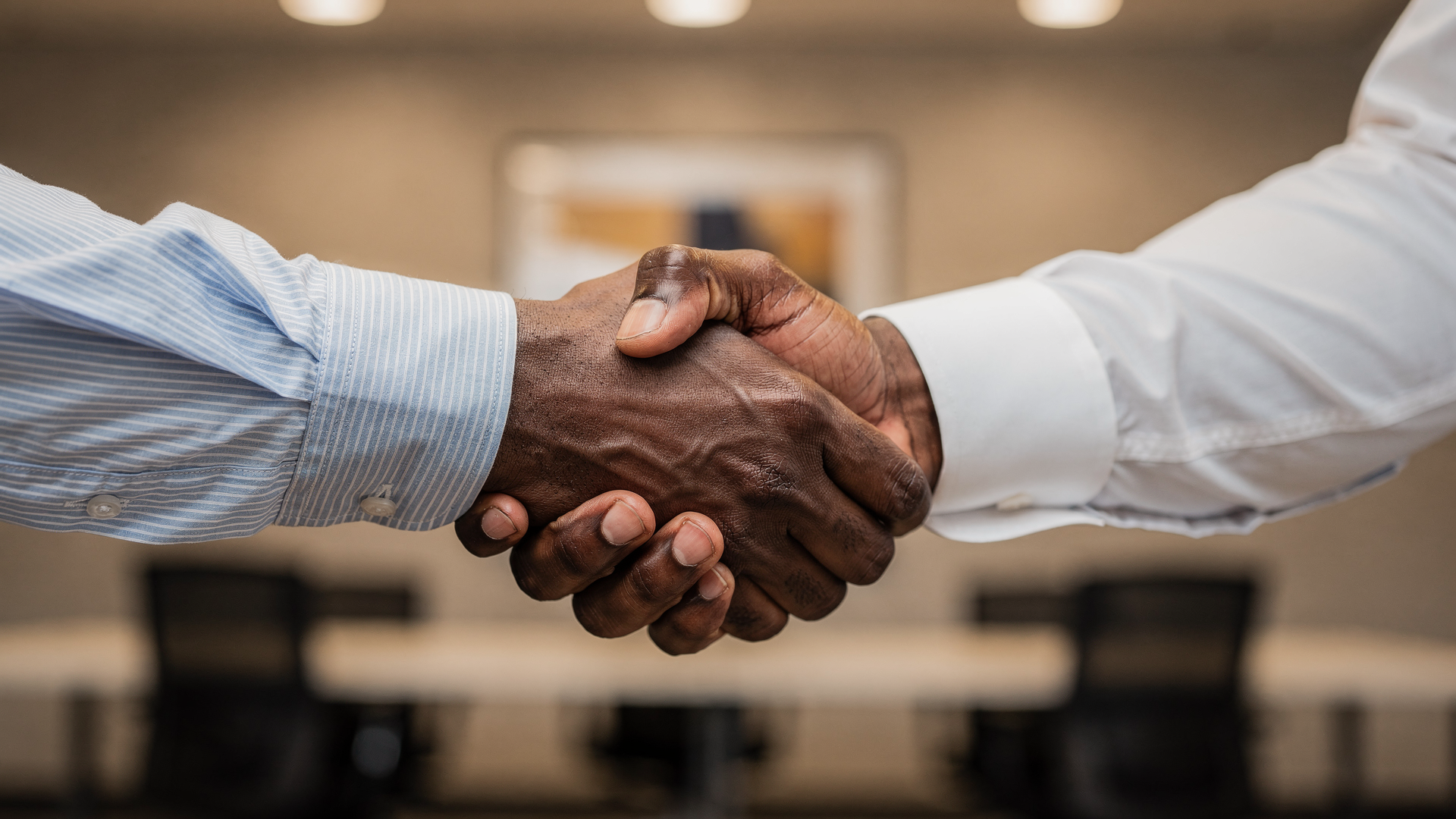  A close-up, medium shot of a handshake between two individuals with dark skin tones. One person wears a long-sleeved, light blue striped dress shirt with the cuff visible, while the other wears a white long-sleeved dress shirt. Their hands are clasp