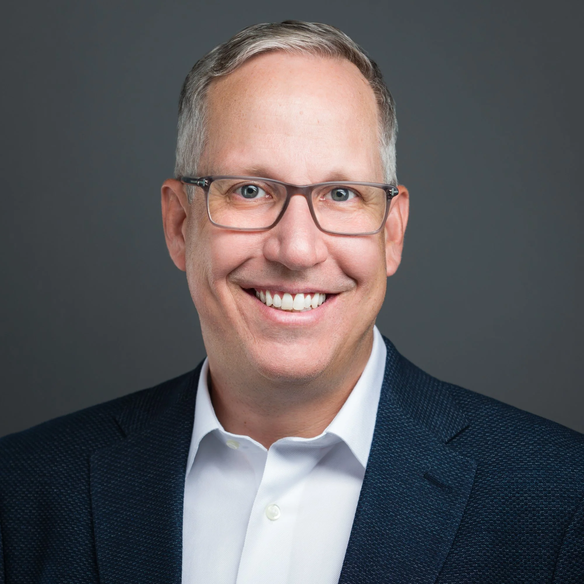 Headshot of a smiling man with glasses, wearing a suit jacket and white shirt, against a dark gray background.