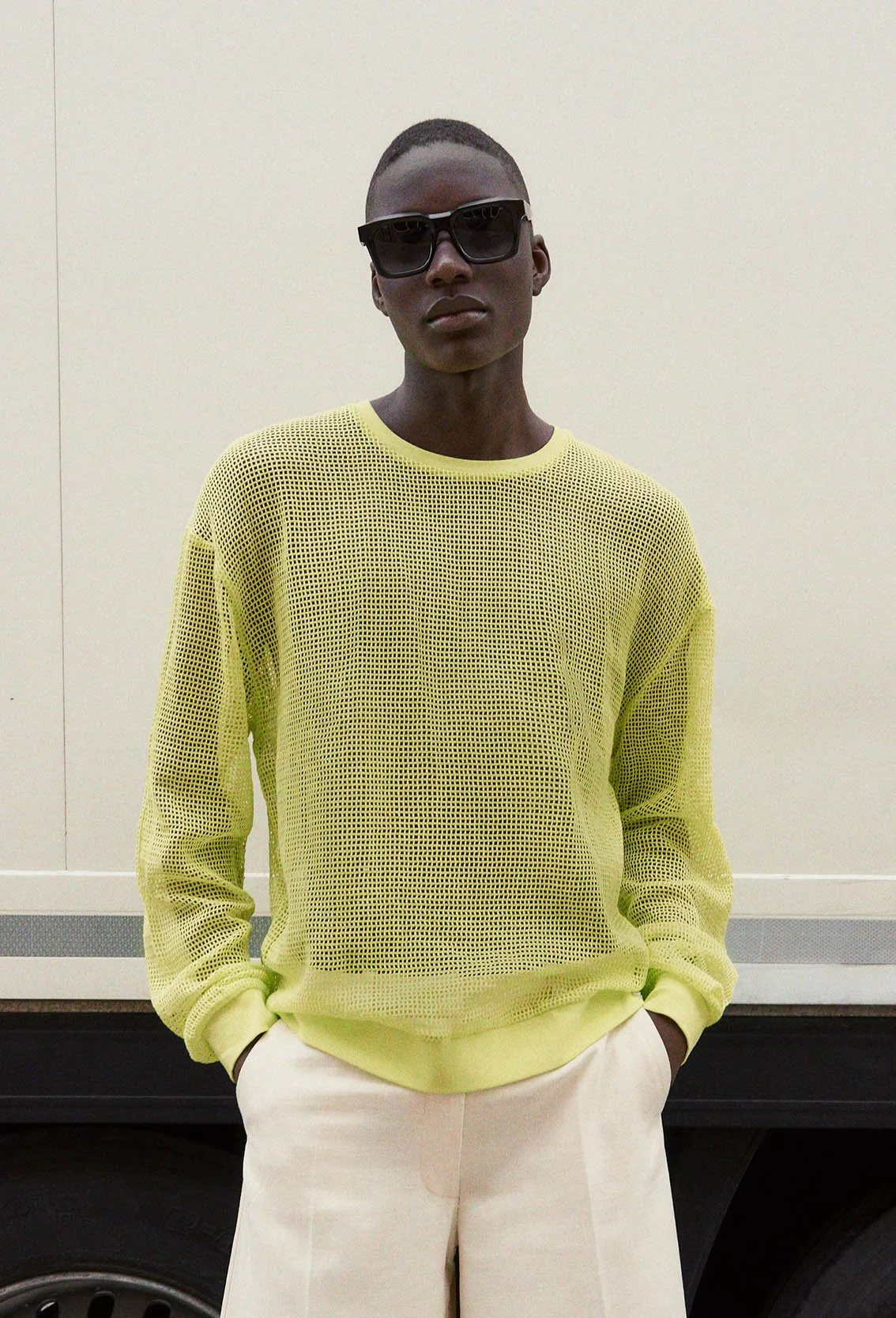 A young man stands in front of a light-colored wall, wearing sunglasses, a yellow mesh long-sleeve shirt, and beige pants.