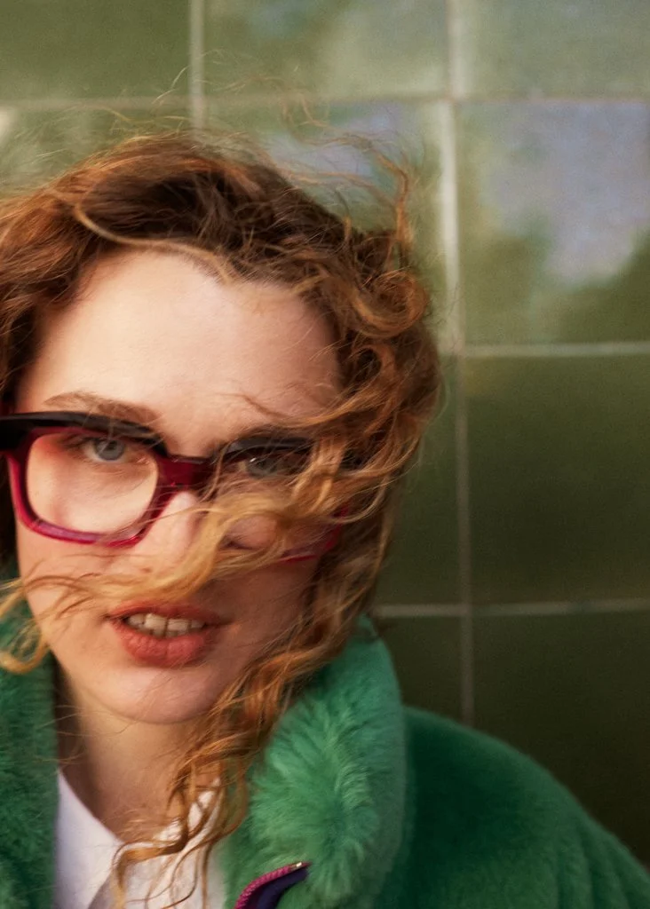 A woman with curly red hair wearing red glasses and a green fuzzy jacket, standing against dark green tiled wall.