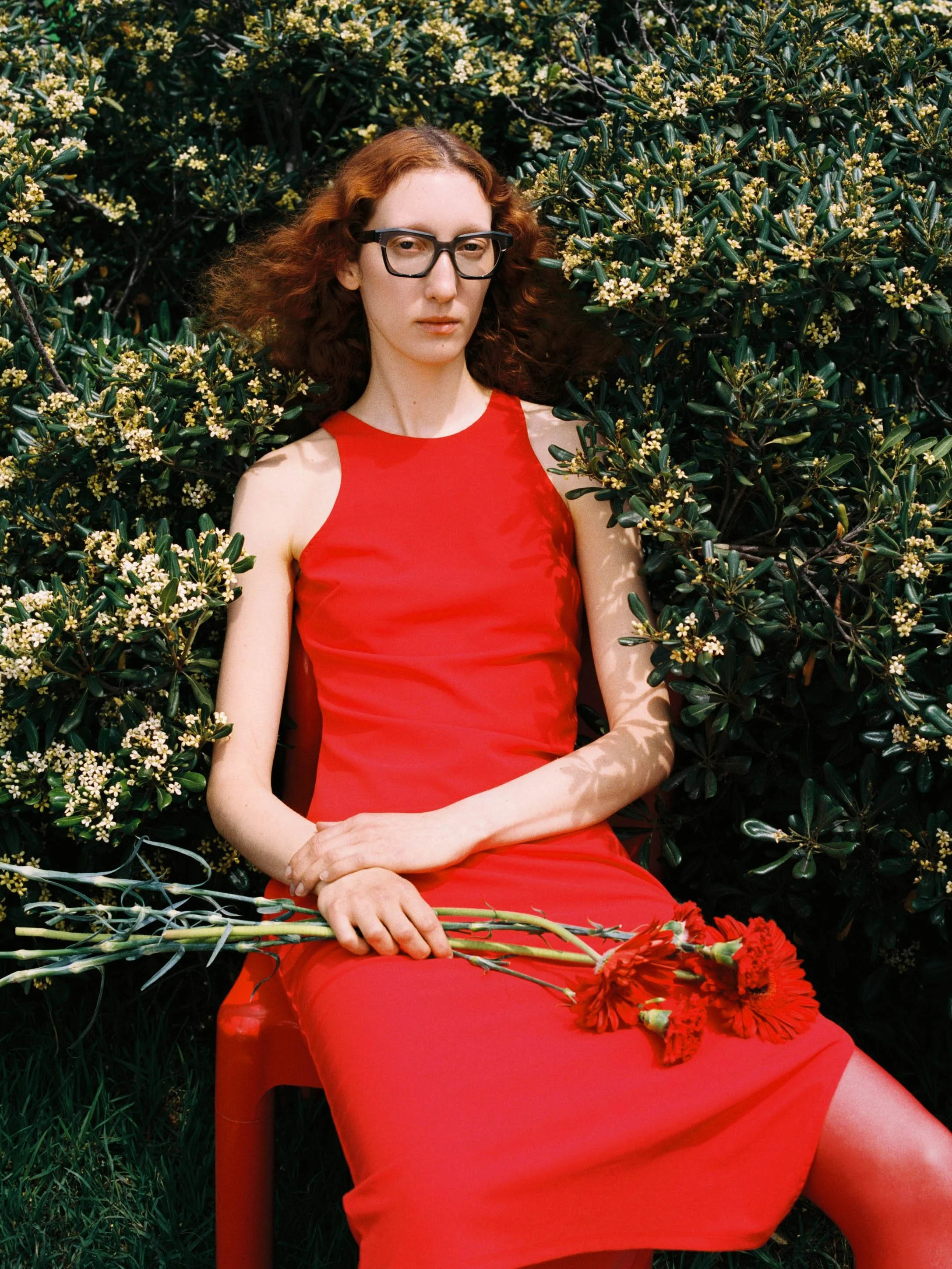 A woman with long red curly hair and glasses wearing a sleeveless red dress, sitting among green bushes with small white flowers, holding red flowers.