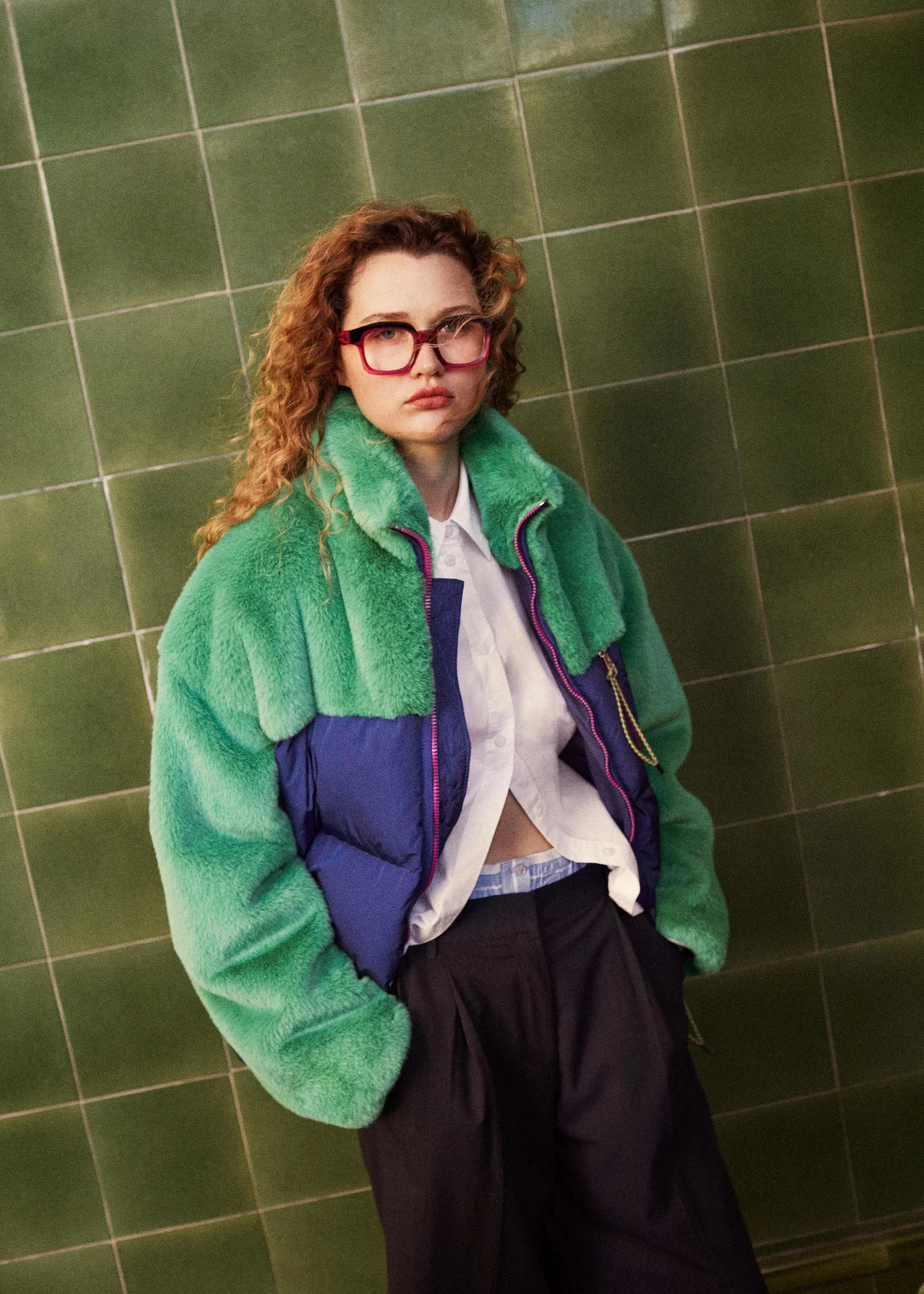 A young woman with curly red hair and glasses wearing a green and purple jacket, white shirt, and dark pants, standing against a green tiled wall.