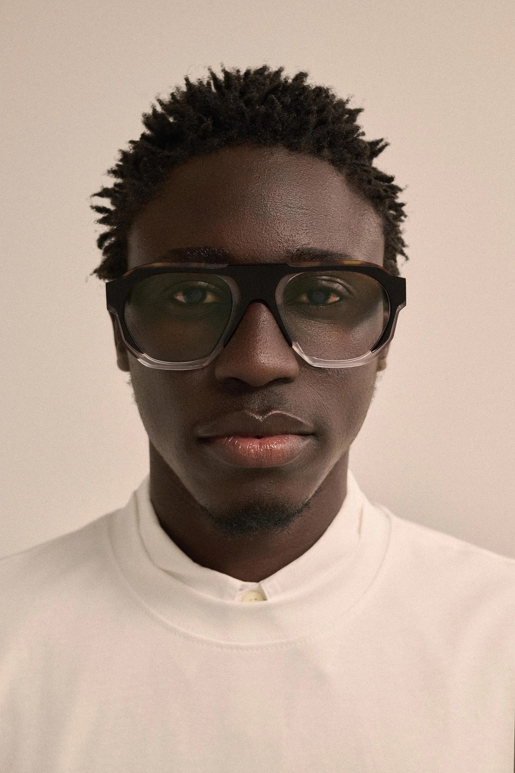Close-up portrait of a person with dark skin, natural textured hair, wearing large black sunglasses and a white shirt with a buttoned collar.