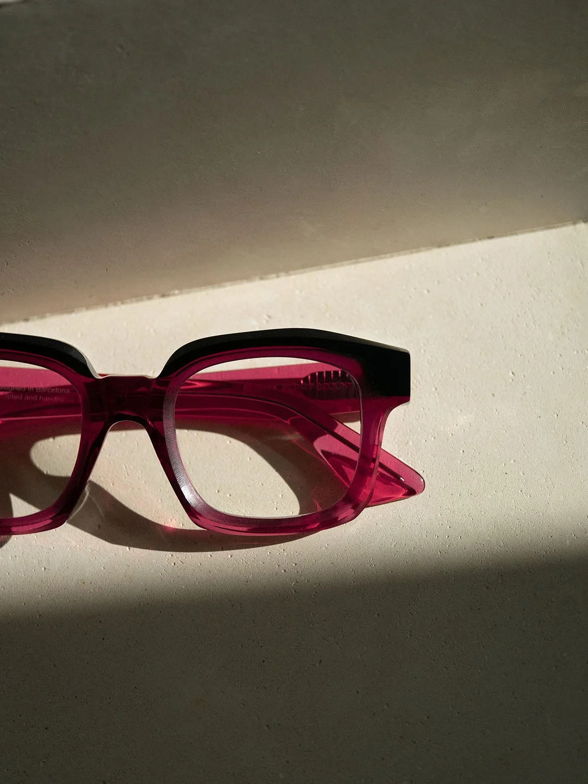 Pink sunglasses with black accents casting a shadow on a light-colored surface with a corner visible in the background.