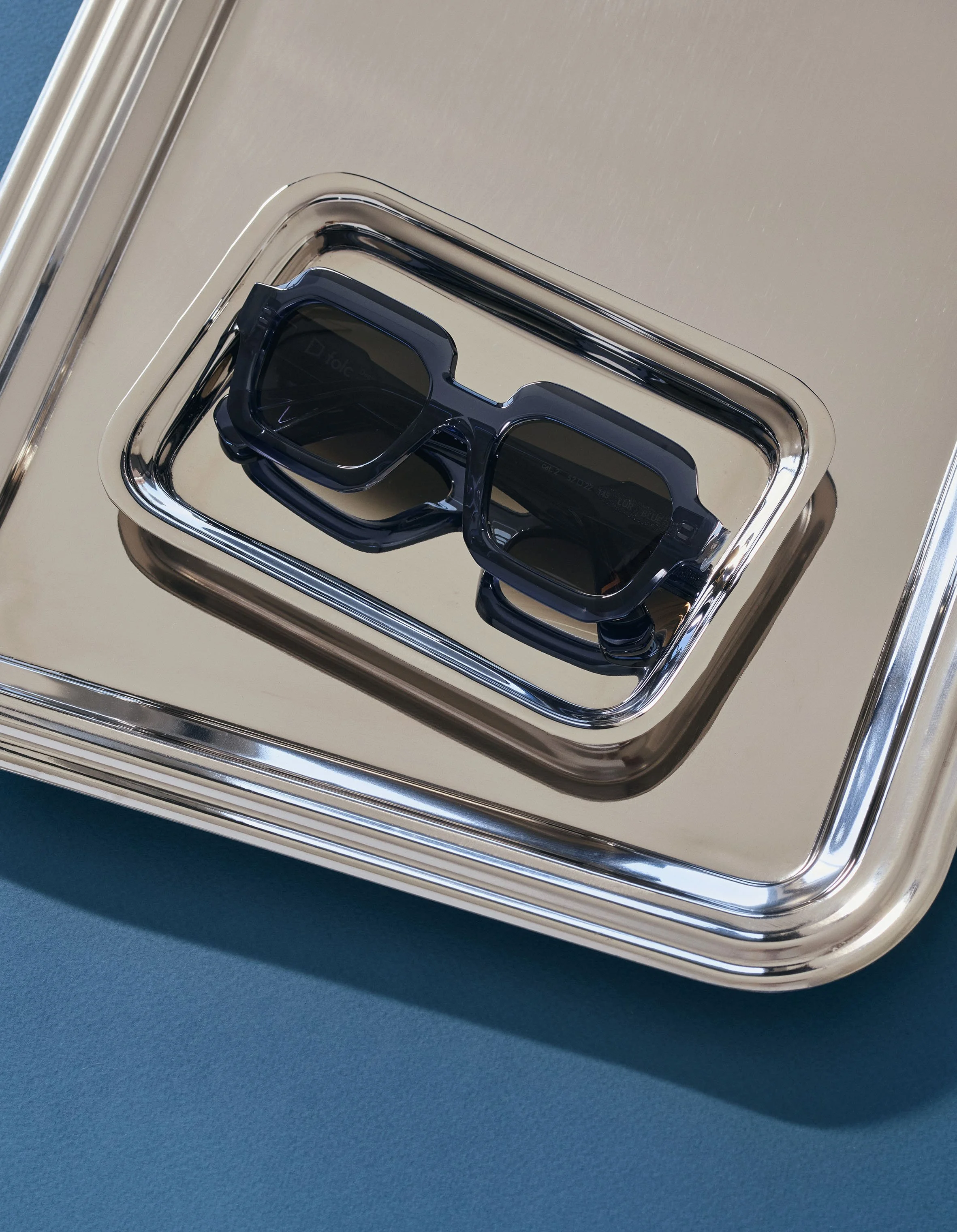 A pair of dark sunglasses resting on a silver tray, placed on a blue surface.