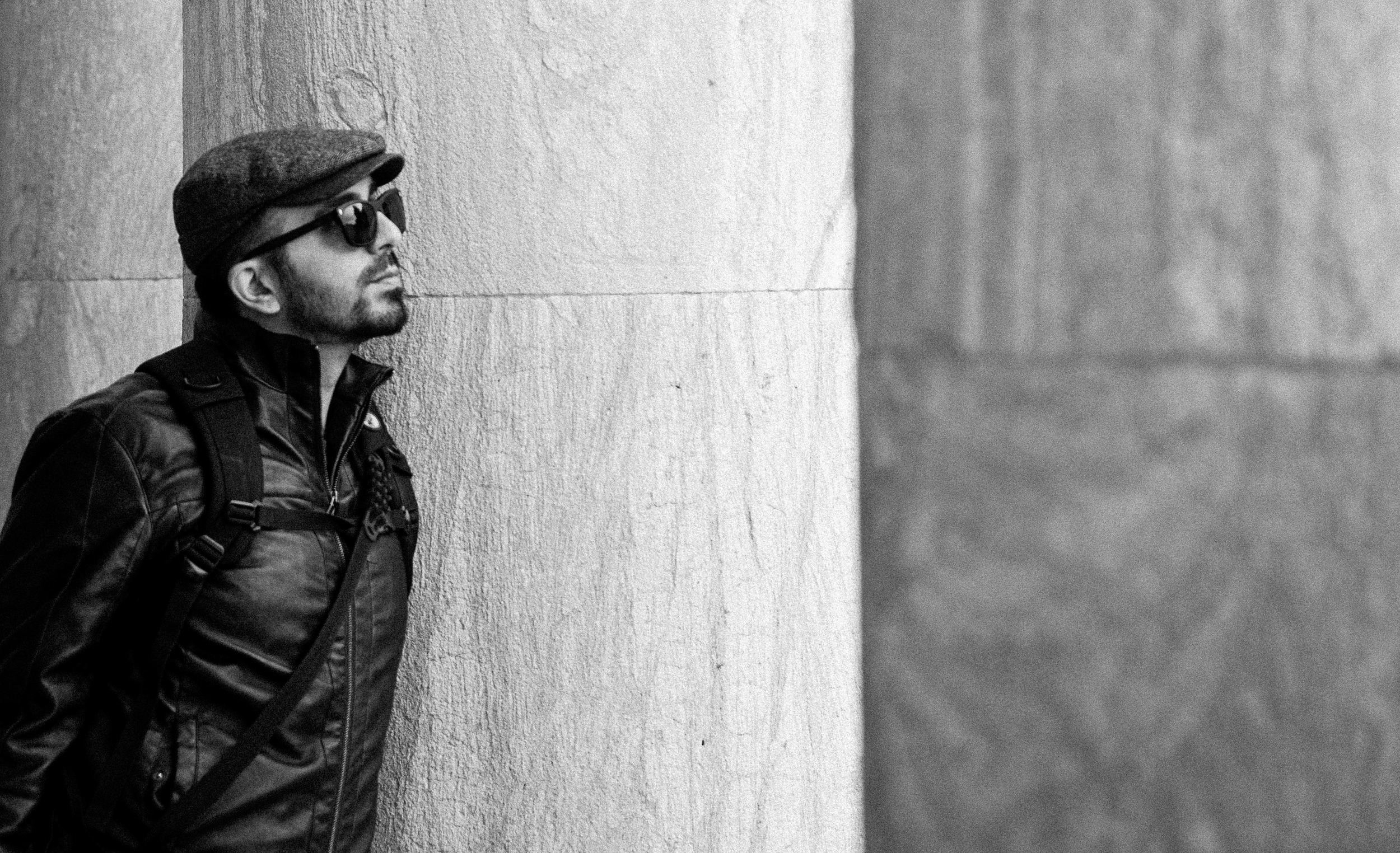 A man wearing a flat cap and sunglasses, dressed in a black leather jacket, leaning against a wall with a serious expression.