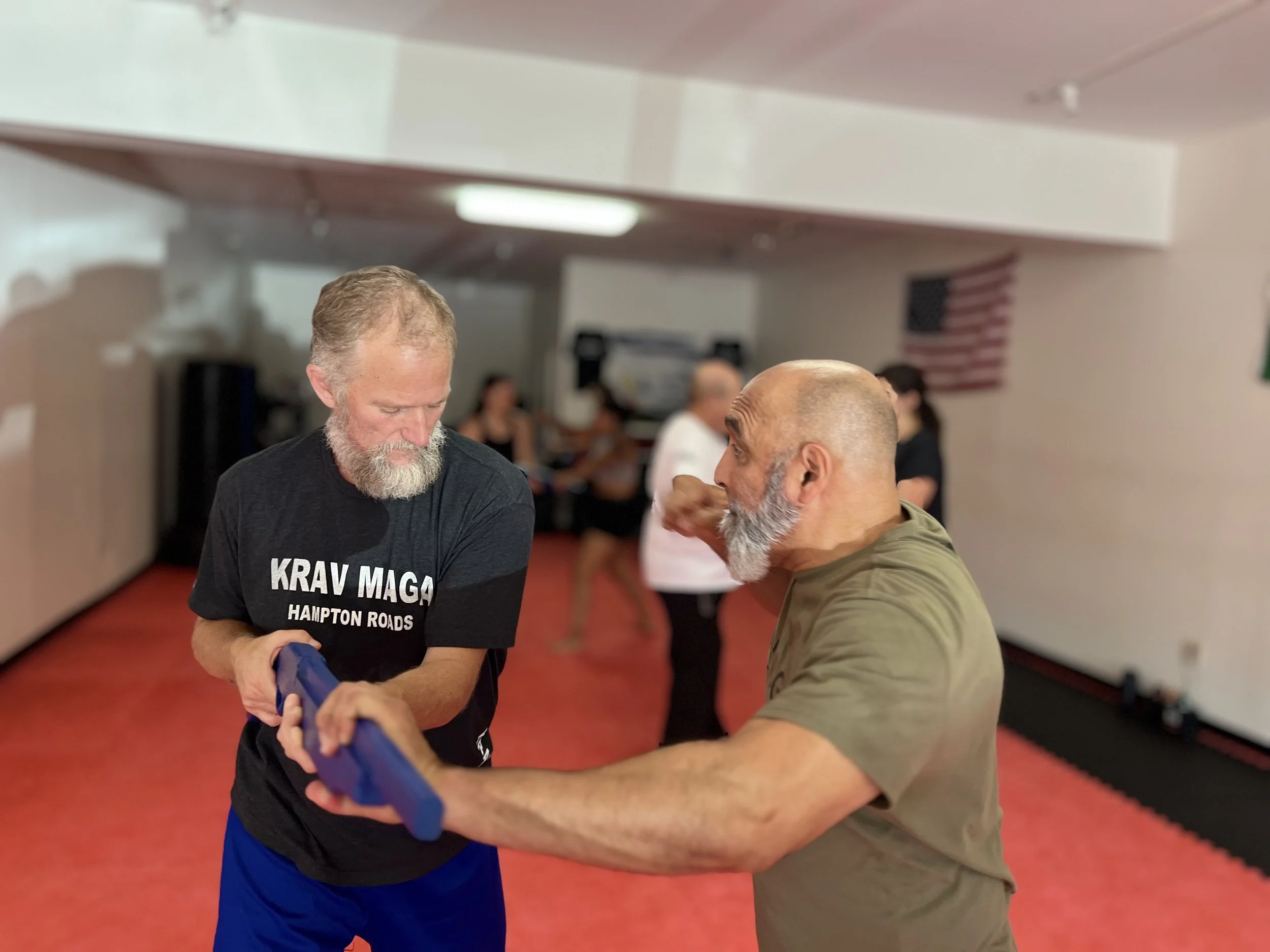 Two older men with gray beards practicing self-defense in a martial arts studio. One man is holding a blue training weapon while the other is guiding him, with an American flag visible in the background.