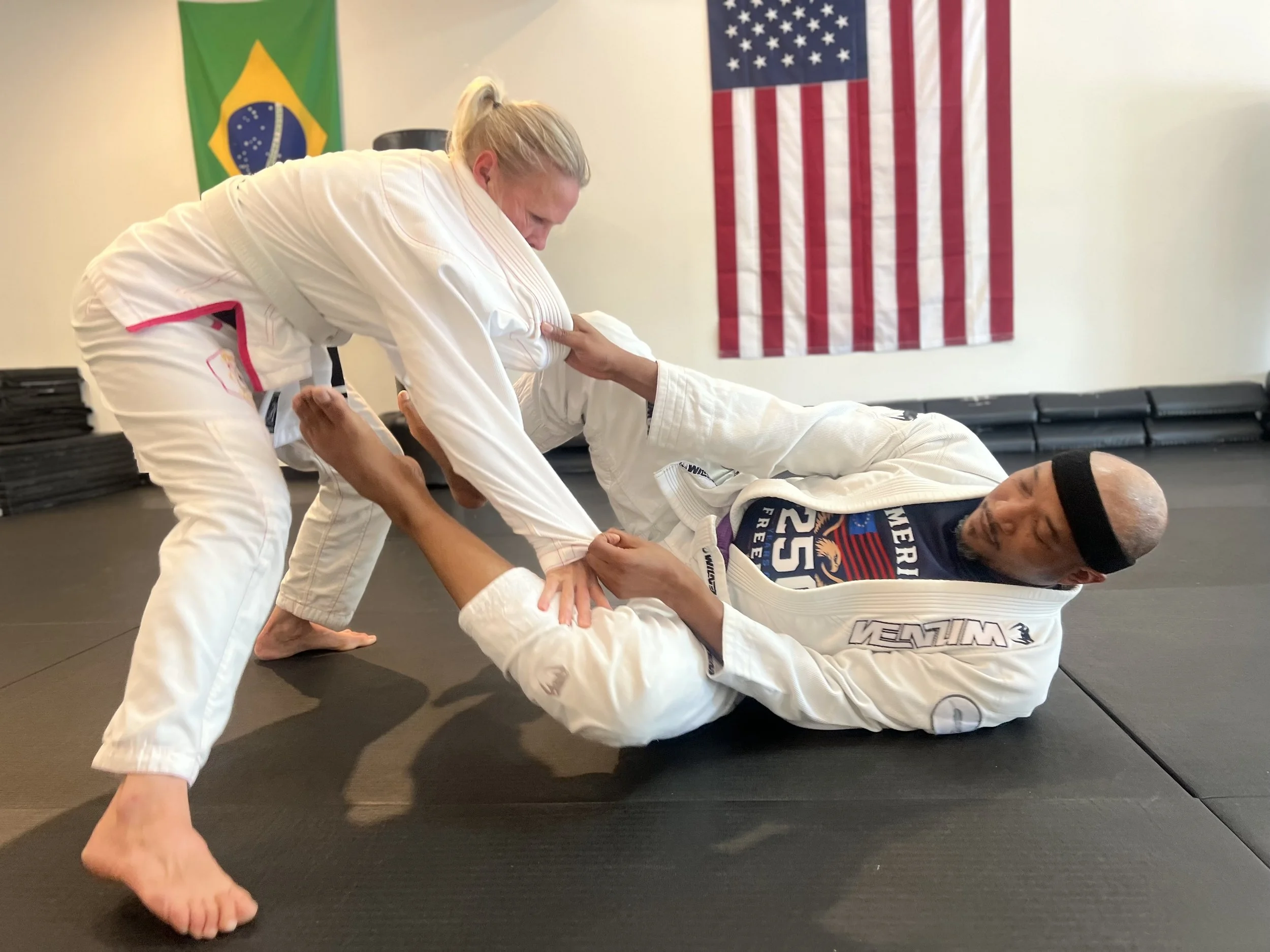 Two pairs of Brazilian Jiu-Jitsu practitioners sparring on black mats, with other students training in the background, American and Brazilian flags hanging on the wall.