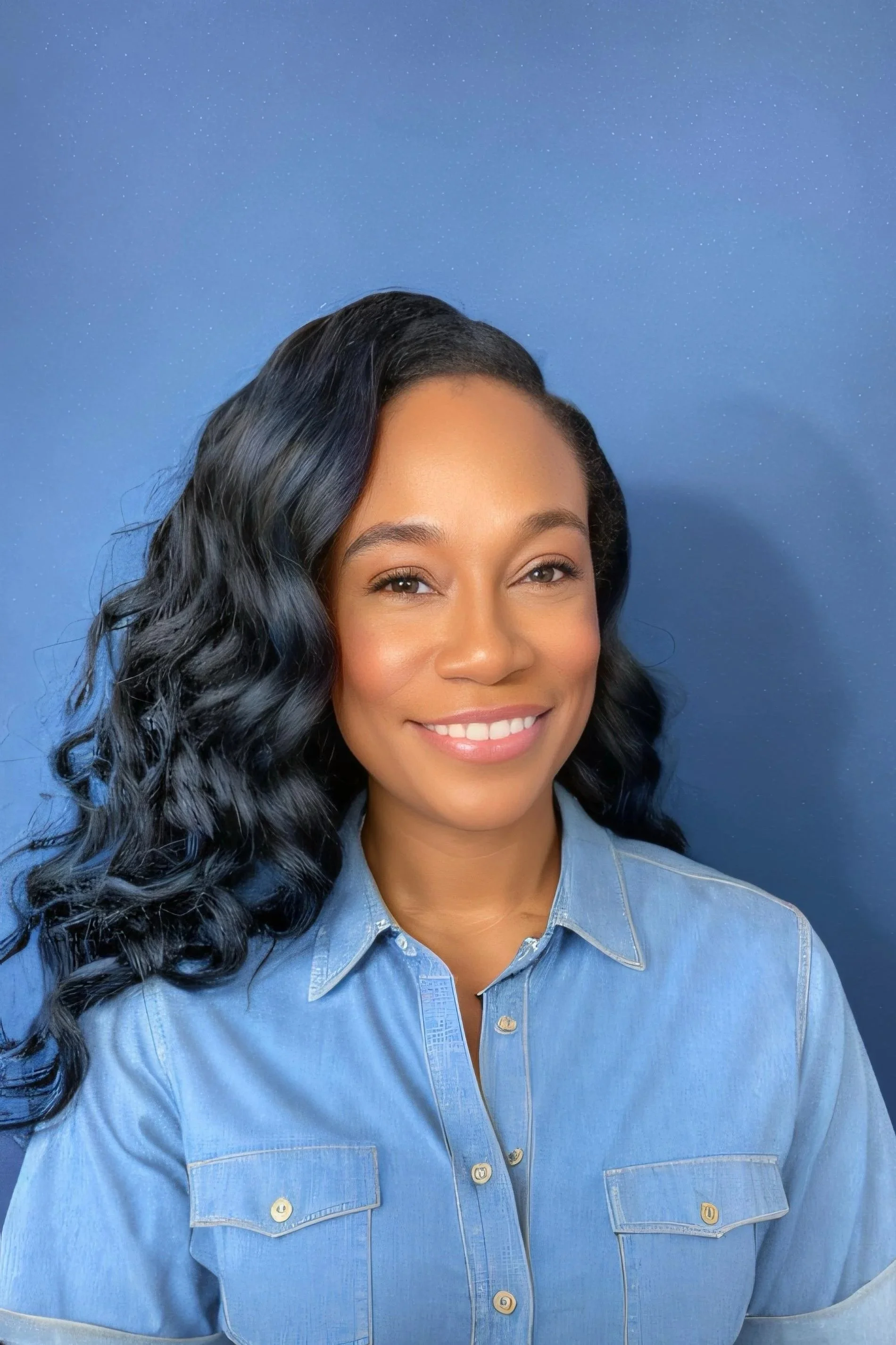 A smiling woman with black, long, wavy hair wearing a light blue denim shirt against a solid blue background.