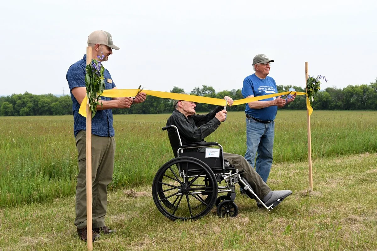 Three people use scissors to cut a ceremonial golden ribbon on the hilltop at Hillside Prairie