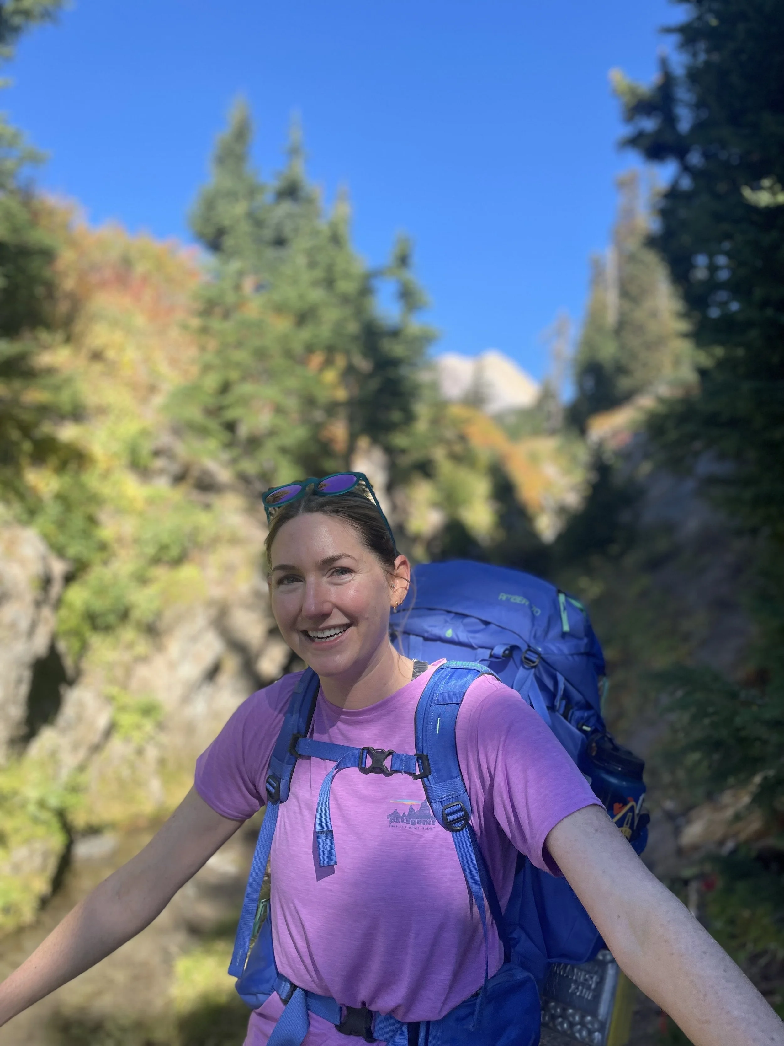 Kady wears a pink shirt, blue backpack, and sunglasses while hiking on a sunny mountainside covered with trees