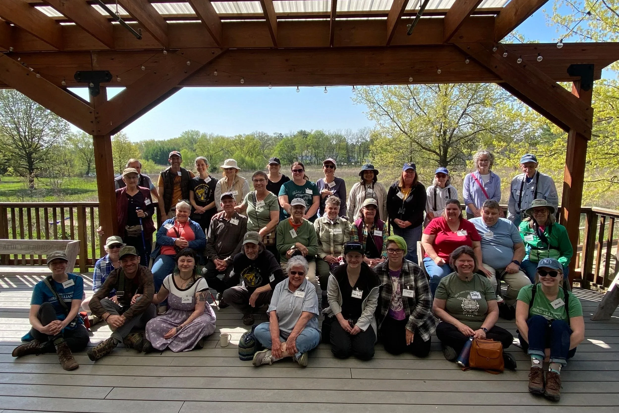 Lots of smiling faces sitting together on the covered outdoor deck at Aldo Leopold Nature Center. The background has lots of trees with green buds.