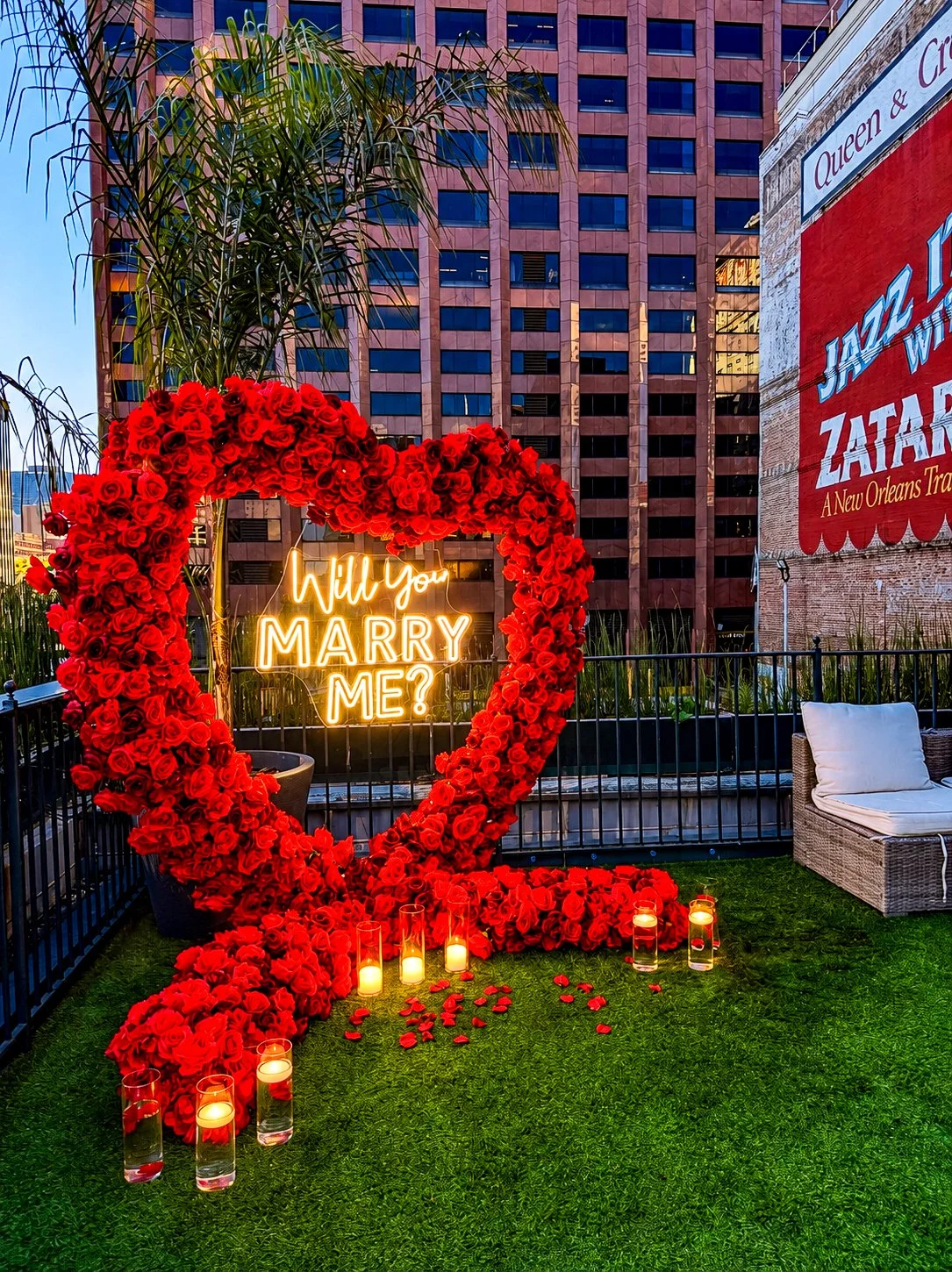 A wedding proposal setup with a large heart-shaped arrangement of red roses, a neon sign inside the heart reads 'Will You Marry Me?', surrounded by candles in glass holders on green grass, with buildings and a billboard in the background.