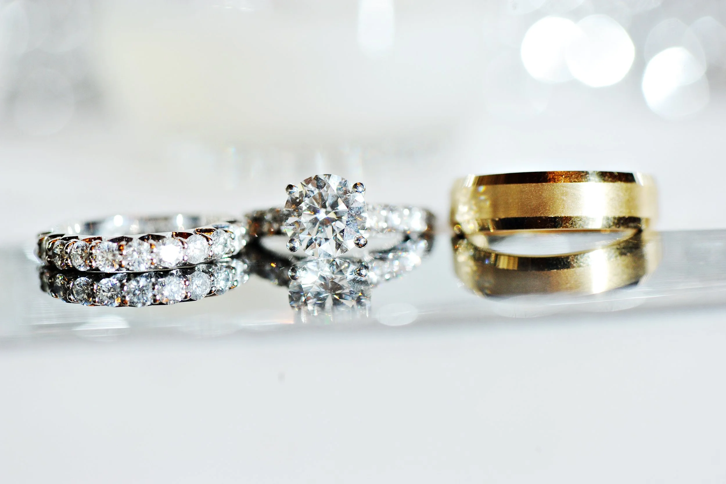 Close-up of three rings: a diamond engagement ring with a large central diamond, a diamond eternity band, and a plain gold wedding band, all reflecting on a shiny surface.