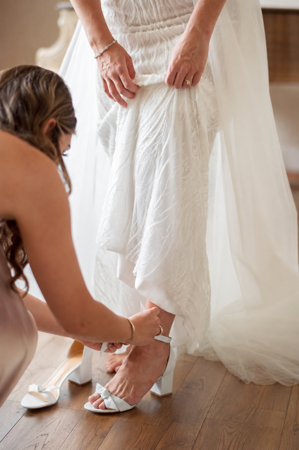 A bride in a white wedding dress standing on a wooden floor while a woman helps her put on white high heels with bows.