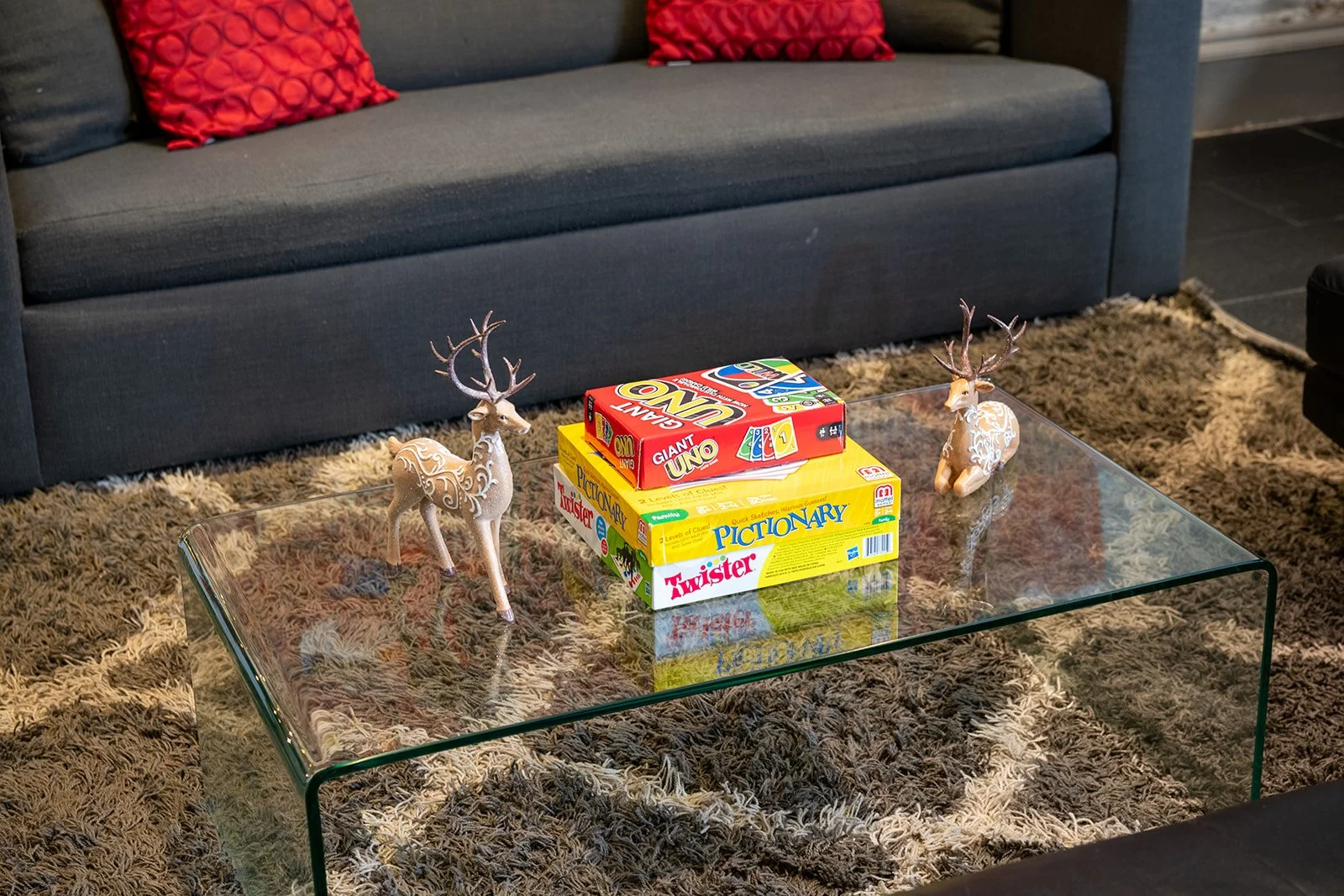 A glass coffee table with two reindeer figurines and three board games inside a living room. The room has a dark gray sofa with red and brown pillows and a brown shaggy rug.