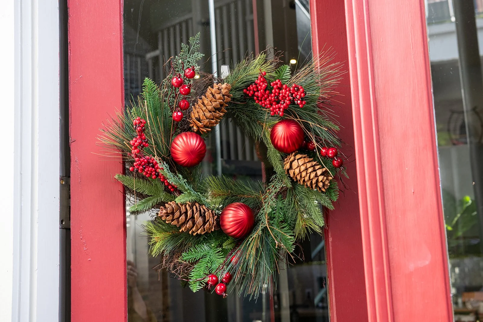 A Christmas wreath decorated with red ornaments, pinecones, red berries, and green pine branches hanging on a red door.