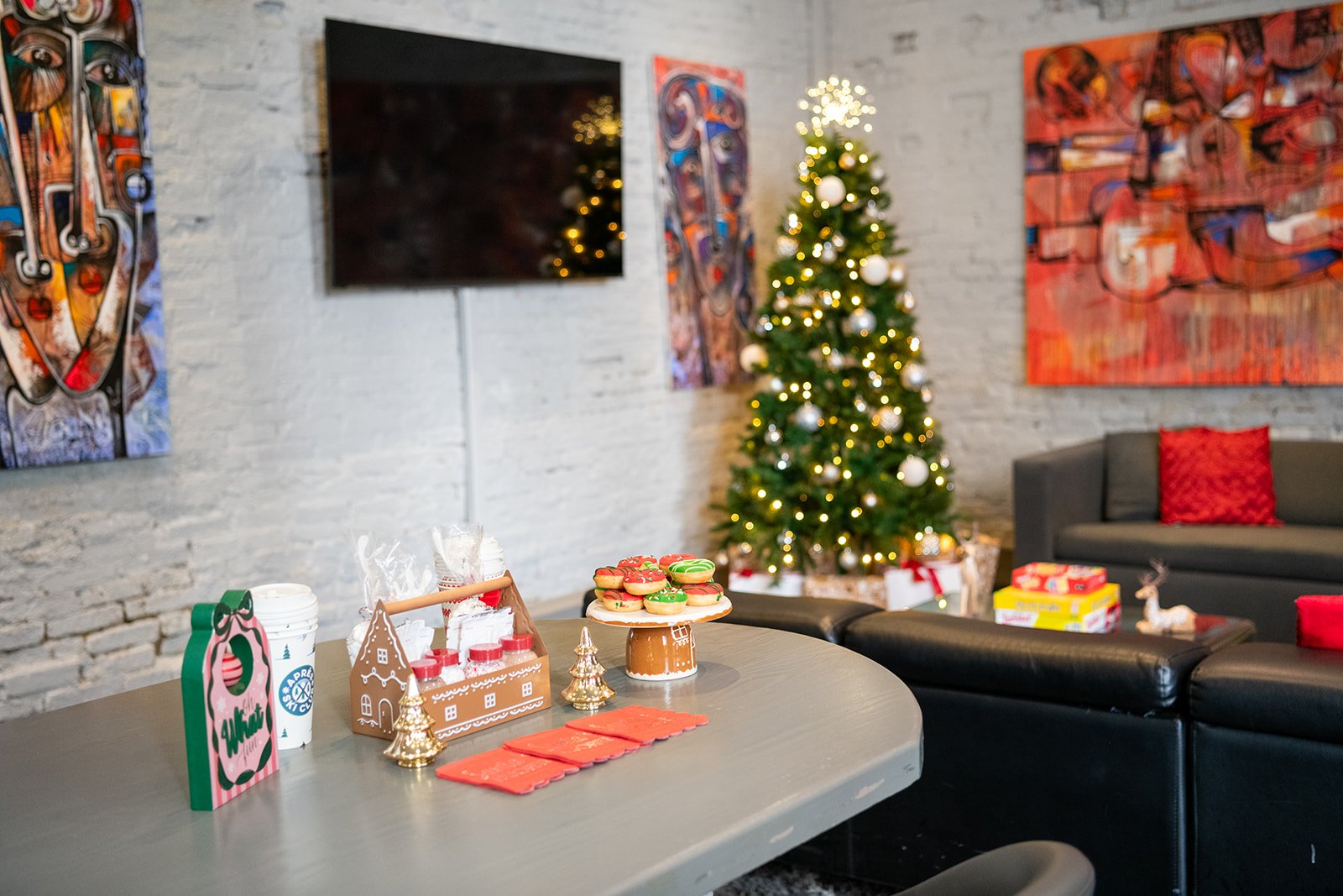 A decorated Christmas tree with white ornaments and string lights in a living room with art on the walls and a grey sofa. A table in the foreground has treats, red napkins, and holiday decorations, with additional presents and decorations visible in the background.