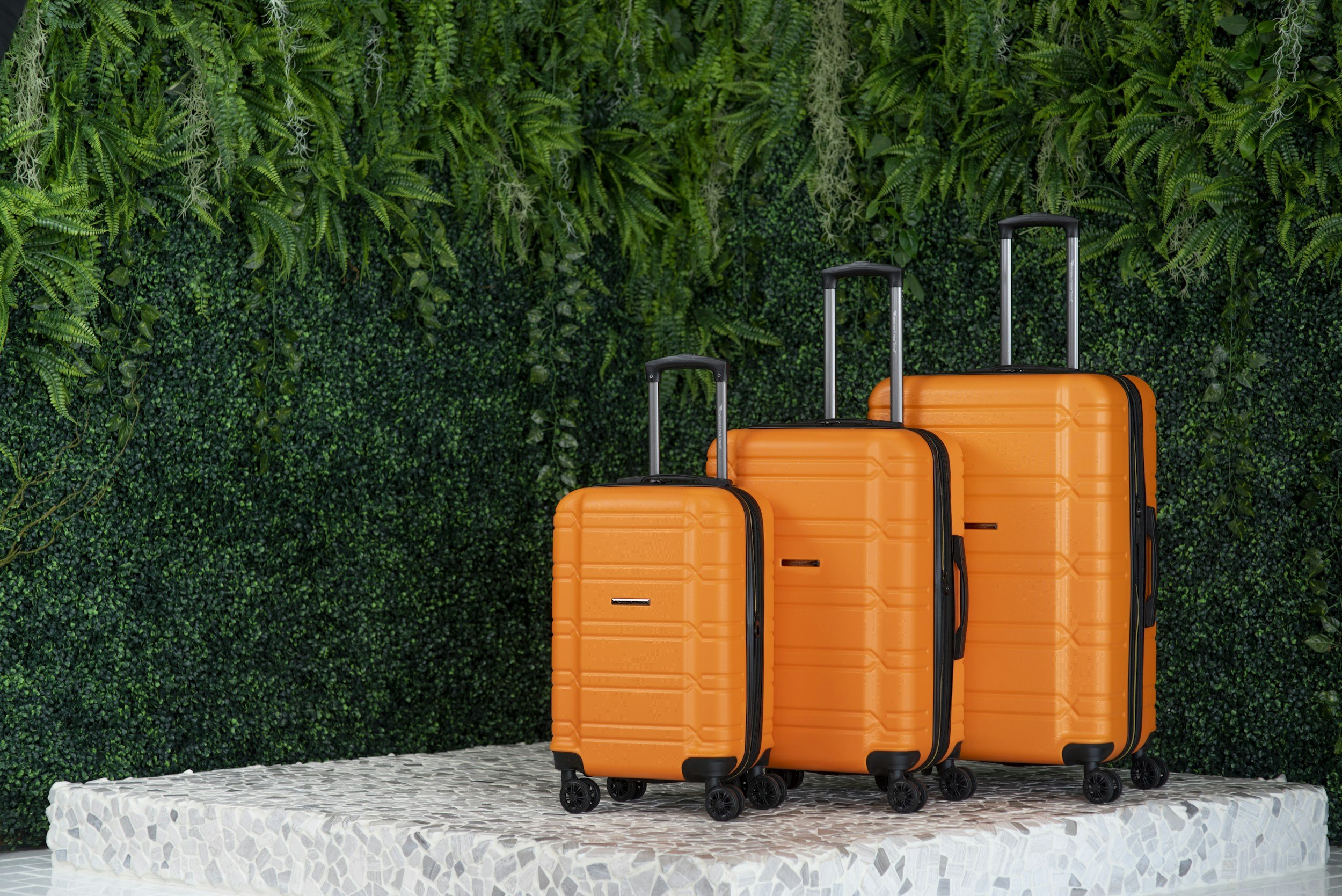 Three orange suitcases of varying sizes with black handles and wheels, placed on a textured white platform in front of a lush green wall with various types of ferns and green foliage.